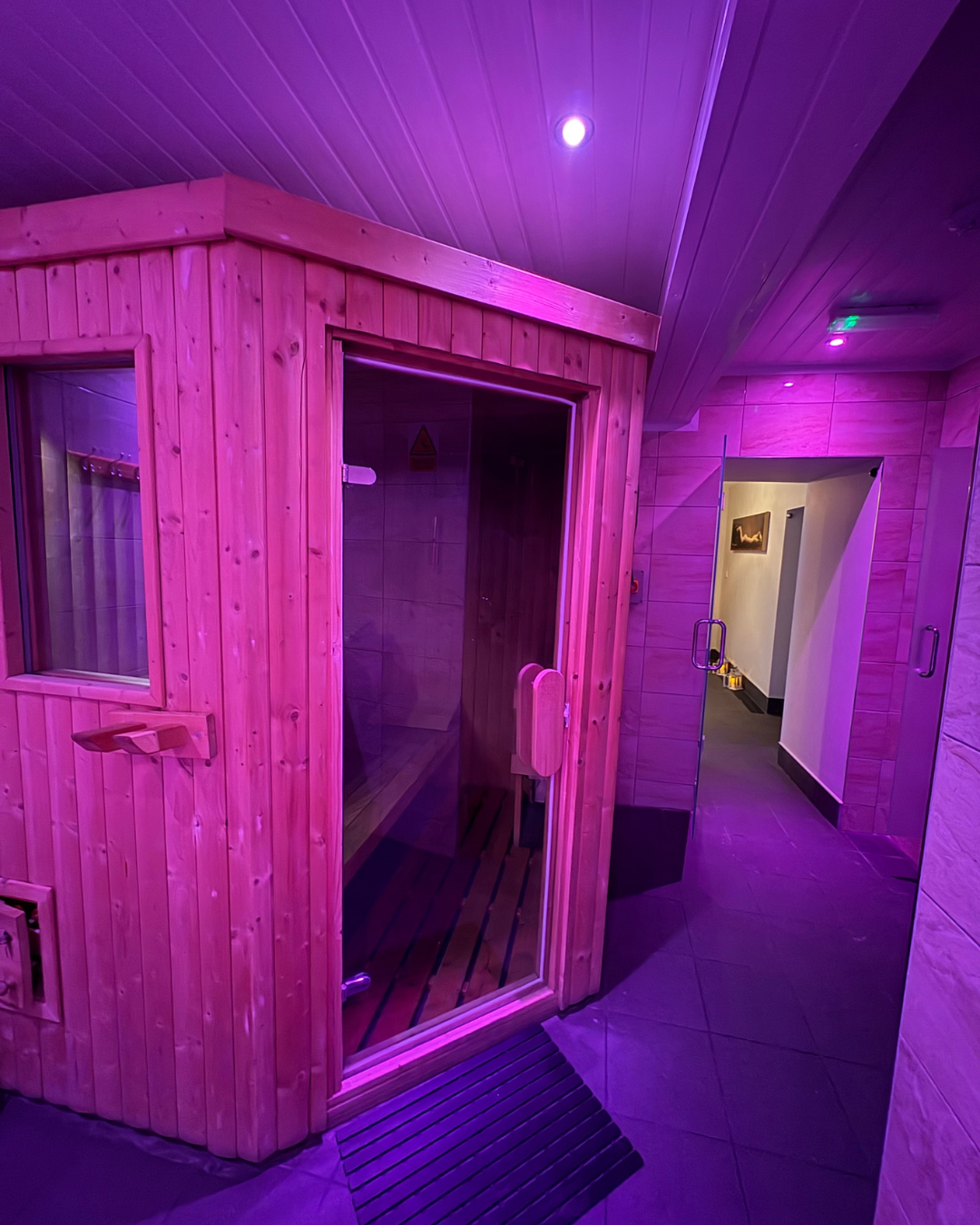 A wooden sauna room illuminated with purple lighting, with a glass door and a small window. The sauna has a wooden bench inside, and the room outside has tiled flooring and a corridor with a light-colored wall.