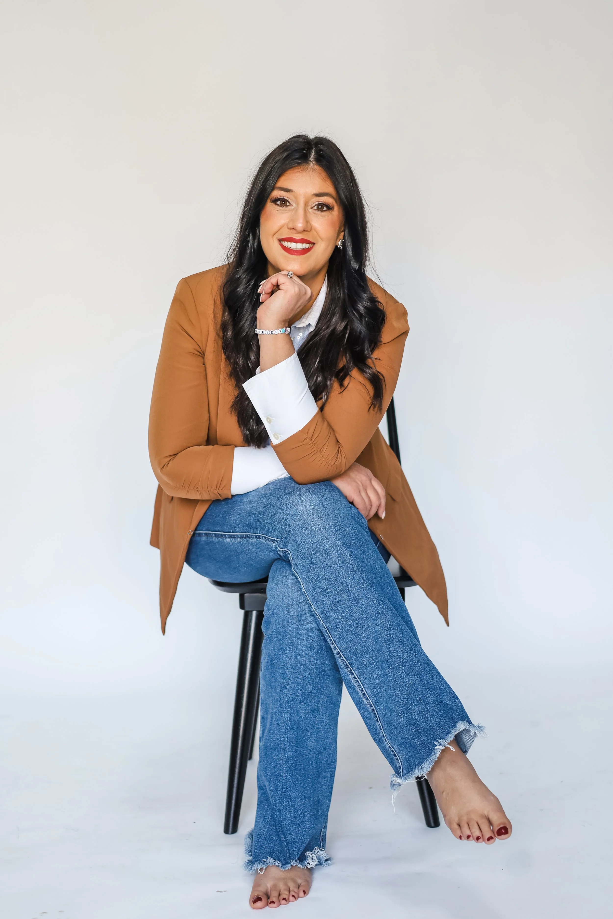 A woman with long black hair, red lipstick, dressed in a brown blazer, white shirt, and ripped blue jeans, sitting on a black chair against a white background, with one hand resting under her chin and smiling at the camera.