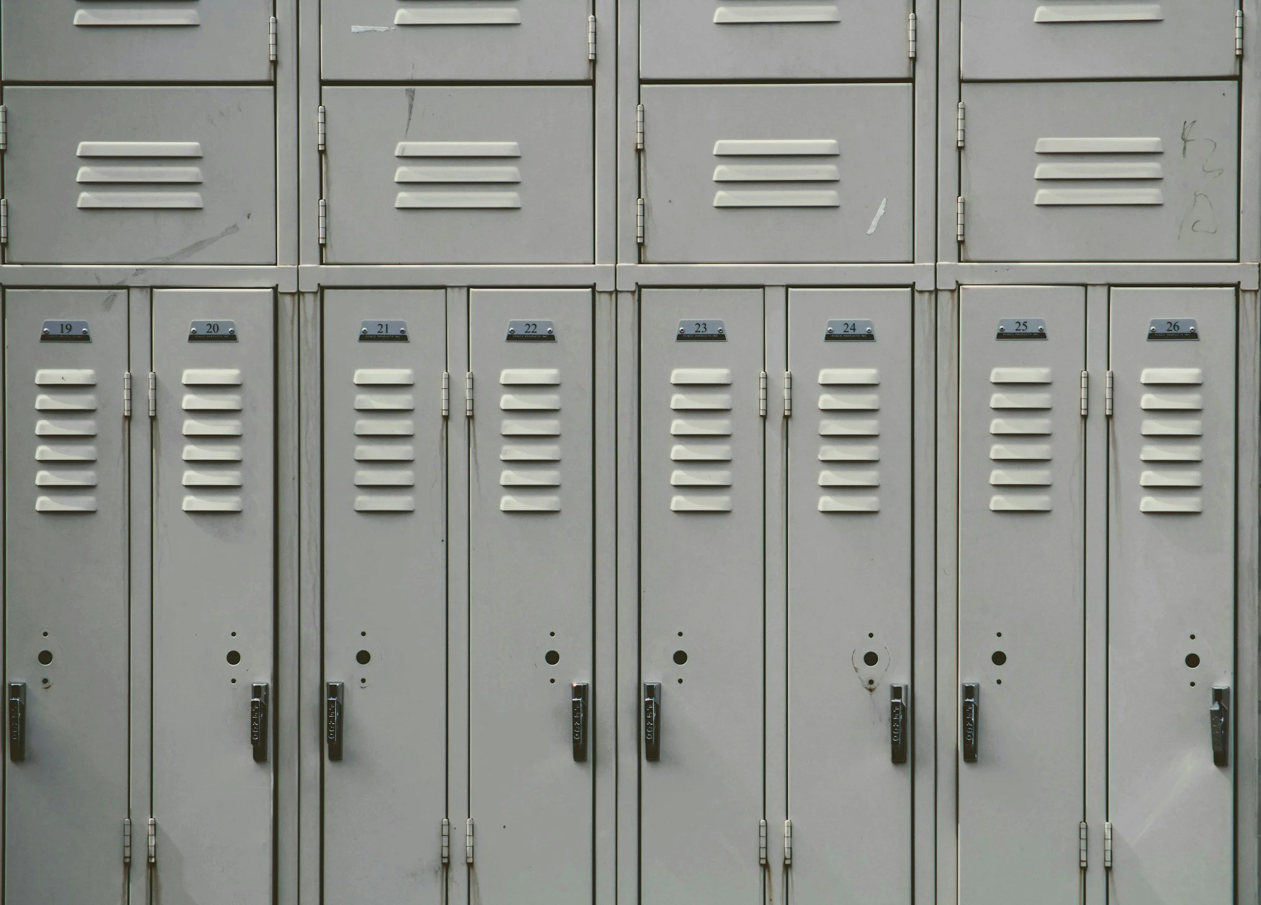 Row of gray lockers with vented doors, some with scratches and graffiti, featuring numbered nameplates from 19 to 26.