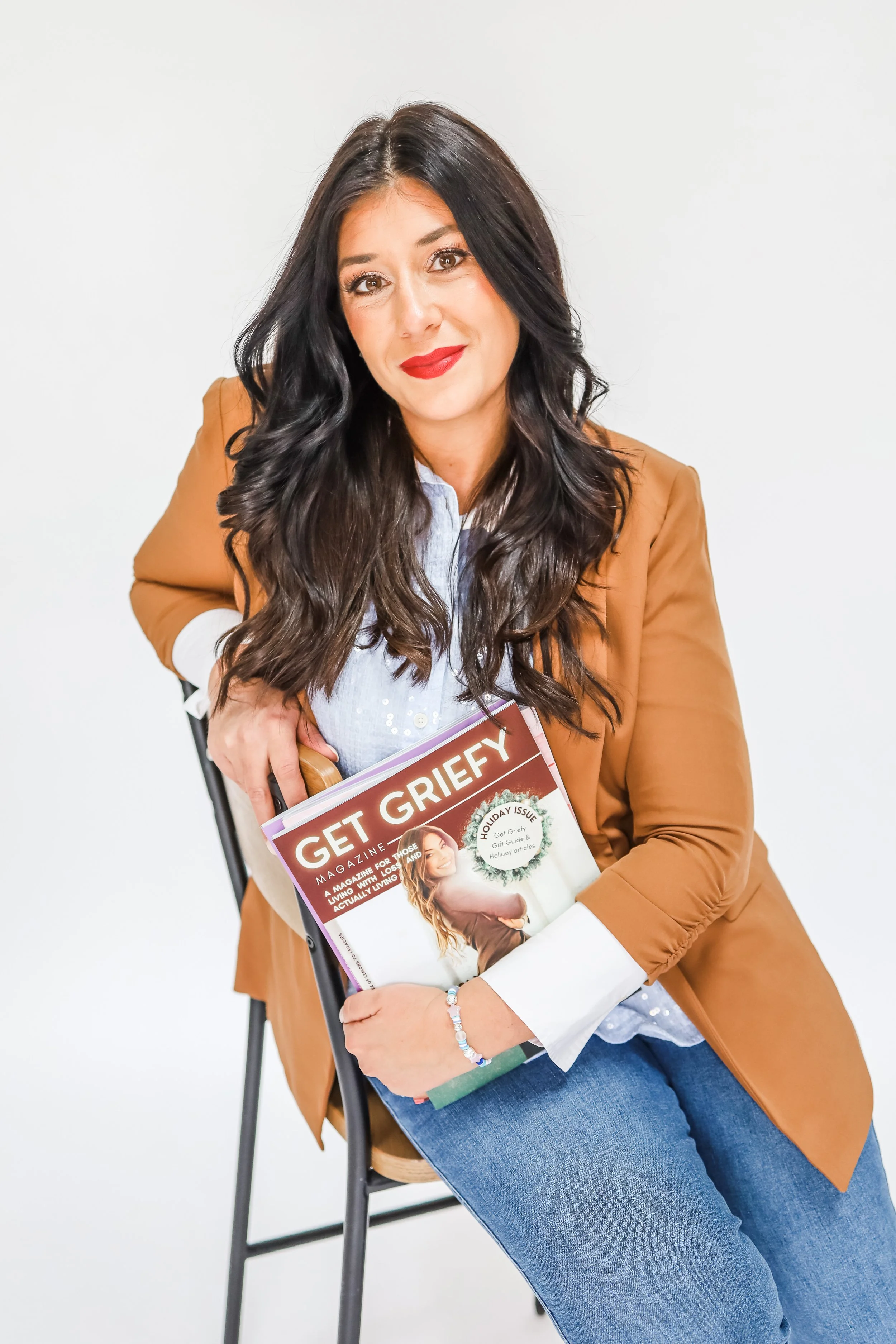 A woman with long dark wavy hair, red lipstick, and makeup, dressed in a light blue blouse and a brown blazer, sitting on a black chair against a plain white background. She is holding a magazine titled 'Get Griefy' with her left hand, and her right hand is resting on her lap. She is smiling slightly and looking directly at the camera.