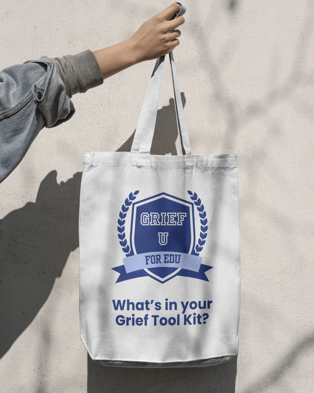 White tote bag with blue text and graphics hanging on a wall, reads 'GRIEF U FOR EDU' with a crest and the question 'What’s in your Grief Tool Kit?'
