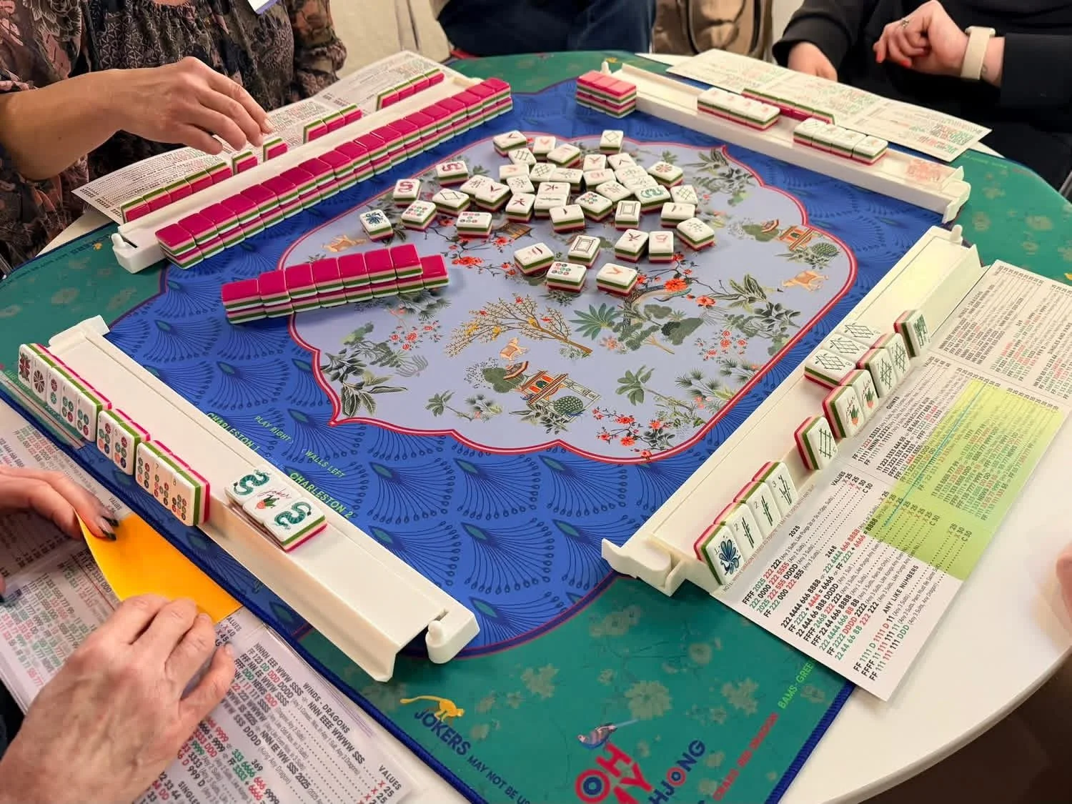 People playing a game of American mahjong with a blue decorative mat, mahjong tiles arranged around the mat, and open instruction booklets and score sheets on the table.