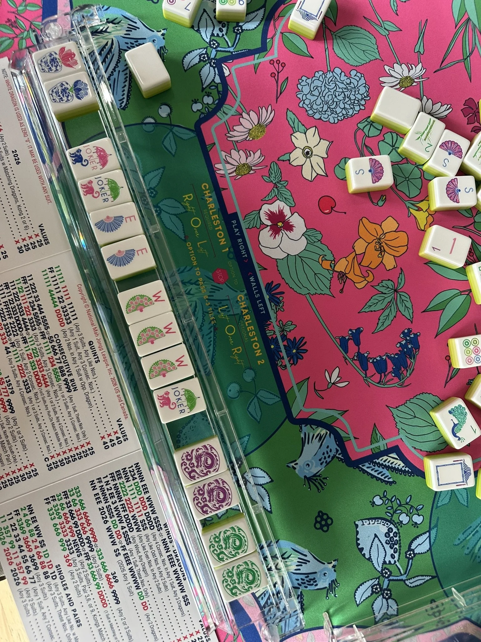 Close-up of an American Mahjong game set with tiles, some of which are arranged on a colorful floral tablecloth. The tiles have various symbols, numbers, and characters.
