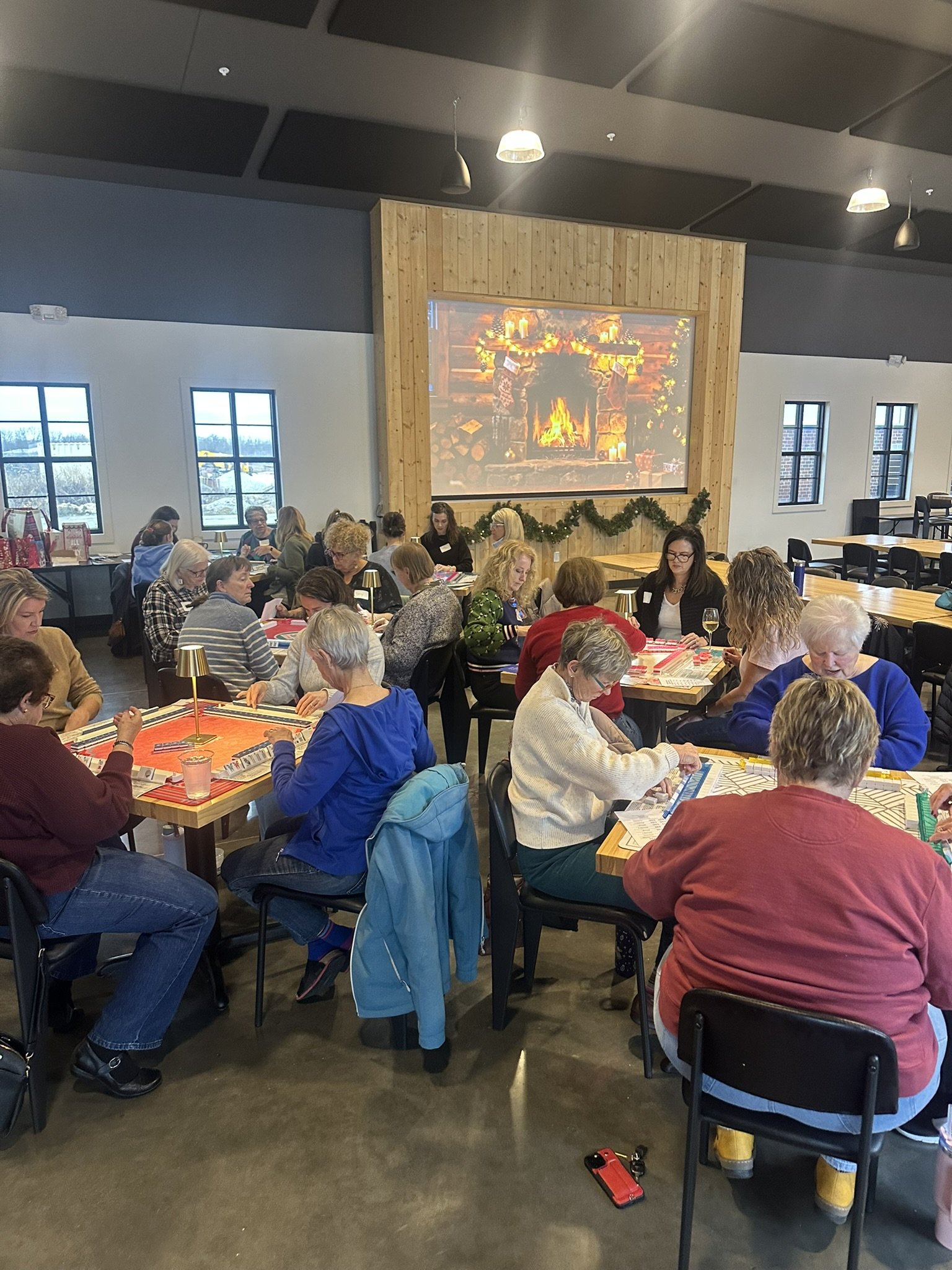 Group of people sitting around tables playing American Mahjong in a festive room with a large fireplace projector screen and holiday decorations.