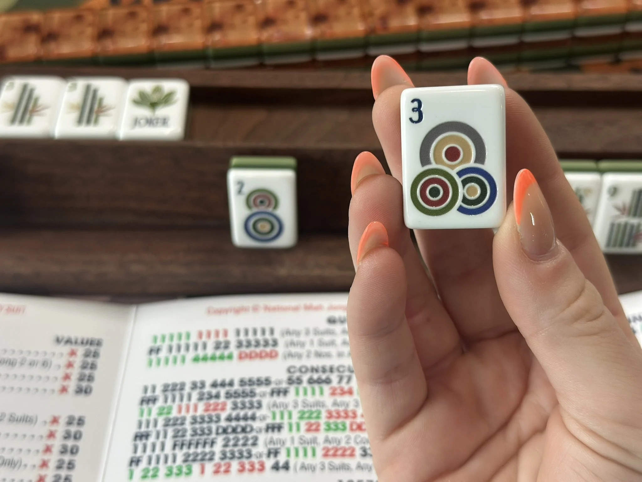 Close-up of a hand holding a dot American Mahjong tile with the number 3 and a multi-colored circular pattern. In the background, other Mahjong tiles and a printed game score sheet are visible.