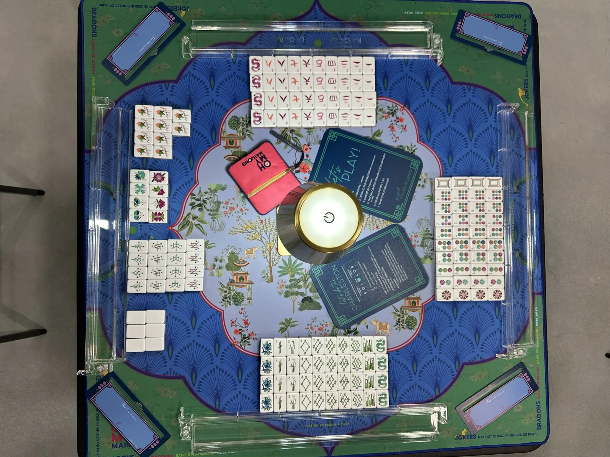 American Mahjong table with various sets of tiles, a pink purse, a round light, and some cards printed with instructions or rules. The table has a colorful floral and animal design, with four corner slots holding additional tiles or cards.