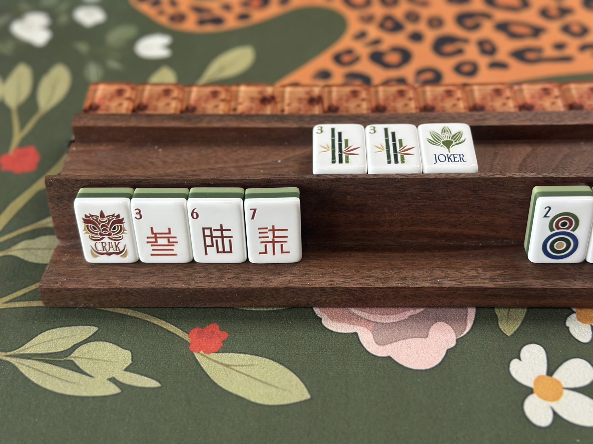 Close-up of American mahjong tiles on a colorful floral tablecloth. The visible tiles include two bamboo suit tiles, grouped together, and a joker tile with a lotus flower design.