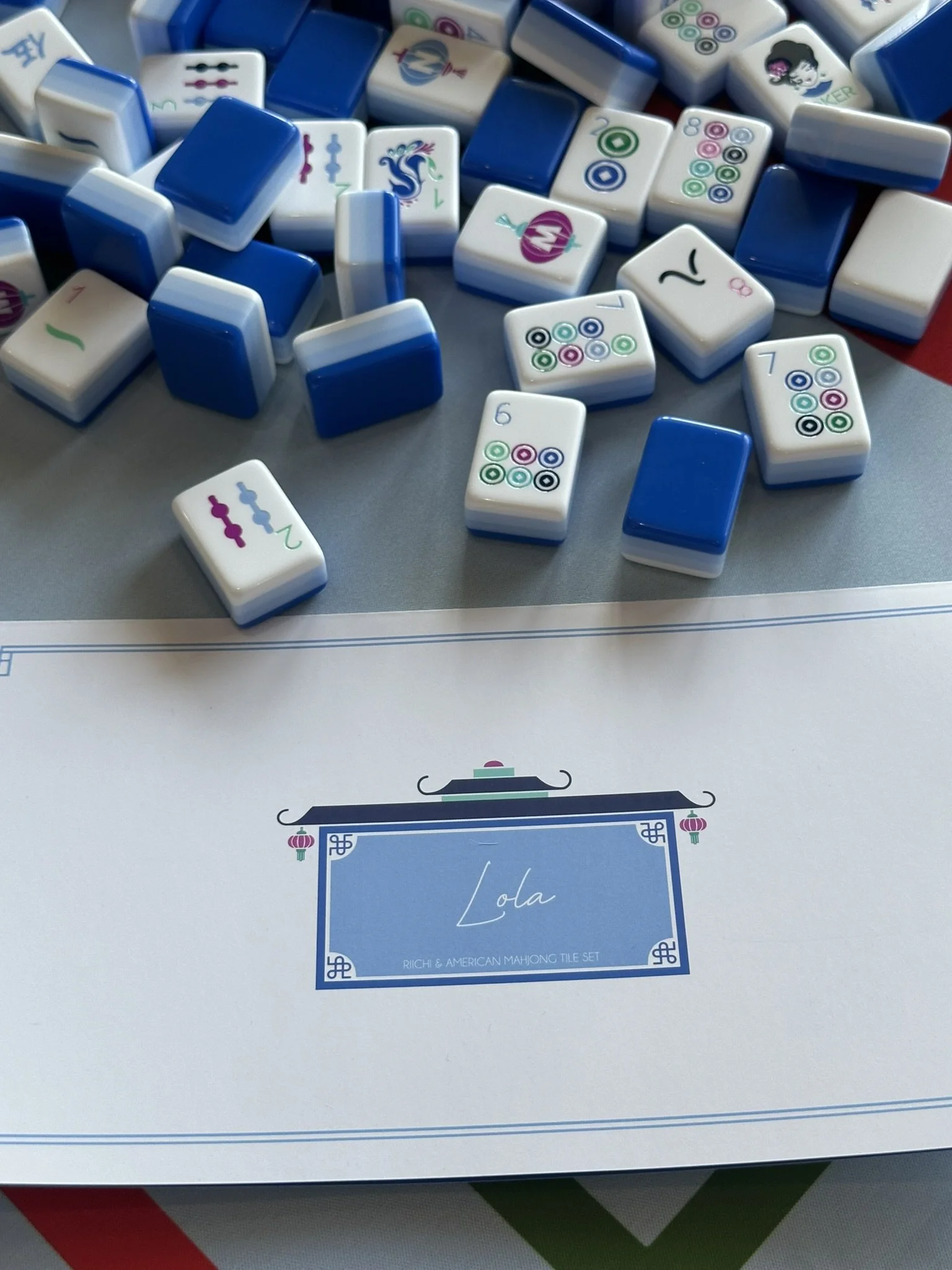 Photo of a set of American Mahjong tiles on top of the box with a blue border design and the text 'Lola' in the center. The tiles are scattered, showing various numbers, symbols, and a dragon design.