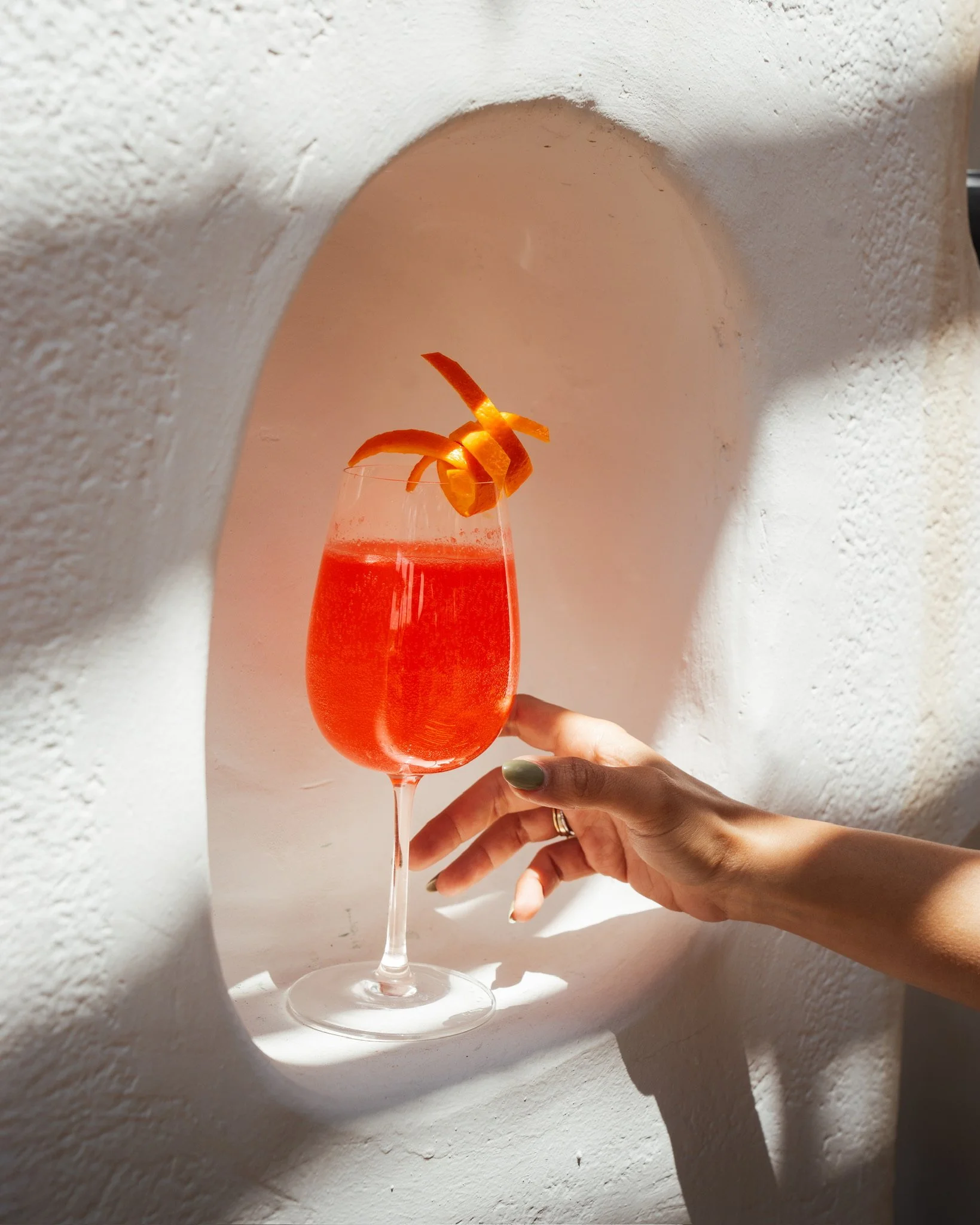 Un verre rose rempli d'une boisson, garni d'une tranche d'orange, tenu par une main avec ongles peints en gris. Fond blanc avec lumière naturelle.