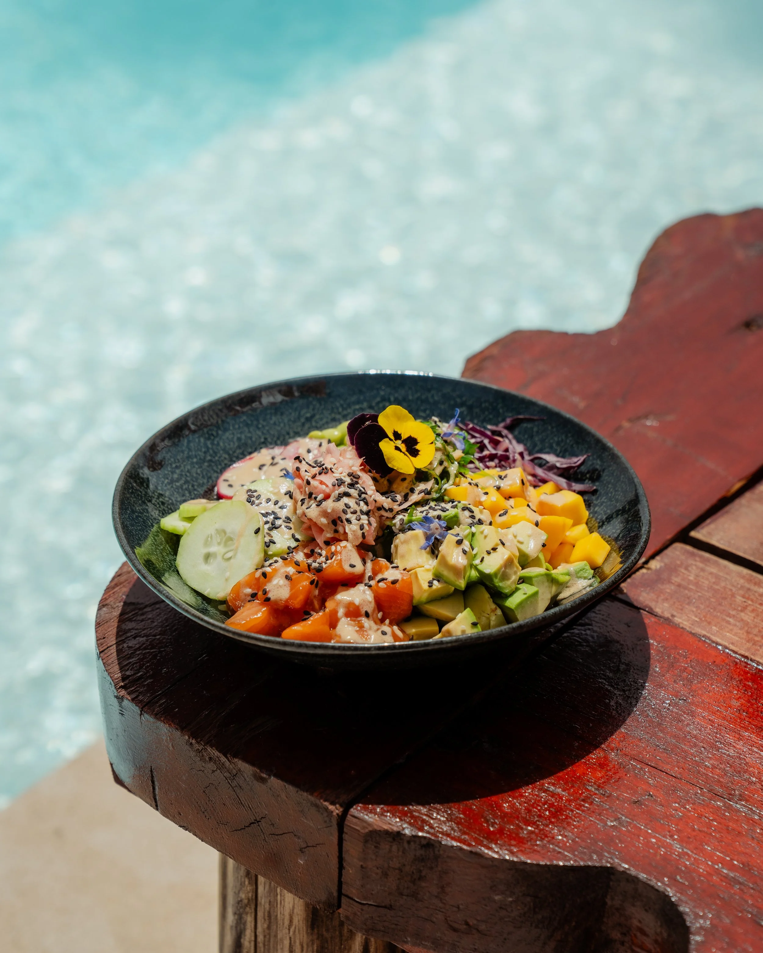 Salade de légumes et poisson dans un bol noir avec des fleurs comestibles, placé sur une table en bois près de la mer.