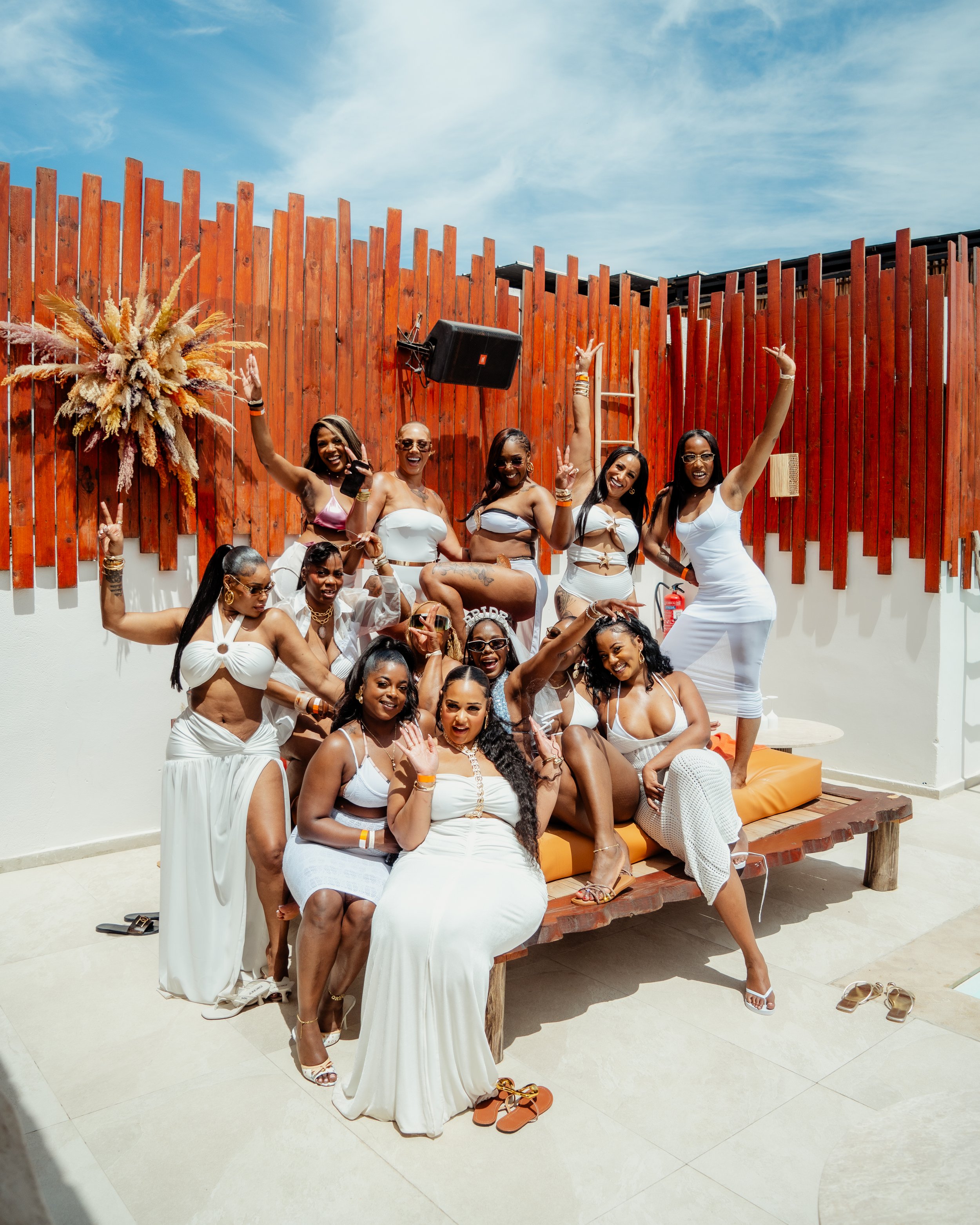 Un groupe de femmes rassemblées, portant principalement des vêtements blancs, posant et souriant devant une clôture en bois rouge avec un ciel bleu en arrière-plan. Certaines femmes portent des accessoires comme des bracelets et des lunettes de soleil, et il y a des chaussures sur le sol.