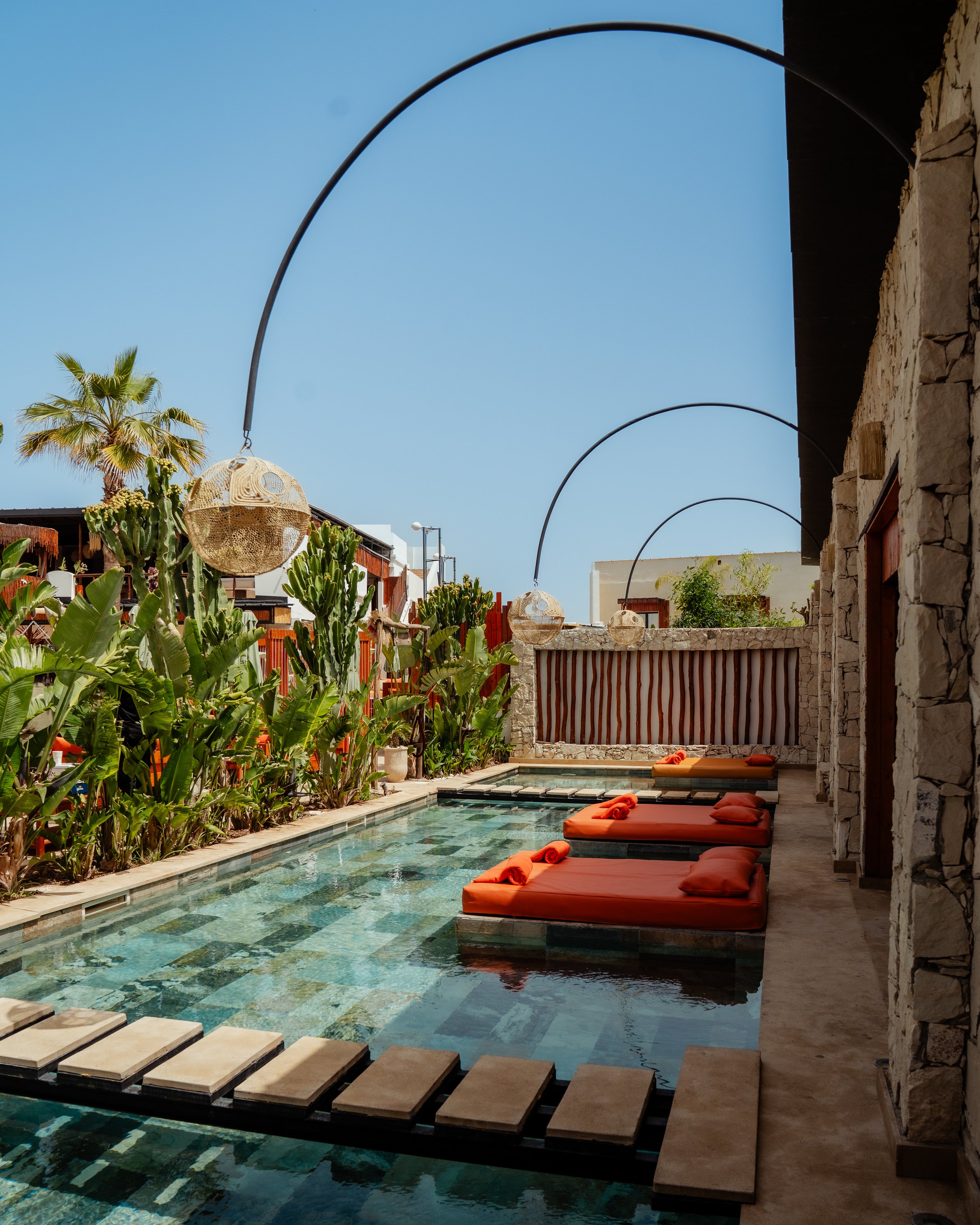 Piscine extérieure avec des coussins orange, des plantes tropicales, des lampes suspendues en osier, et un mur en pierre, avec un ciel bleu au-dessus.