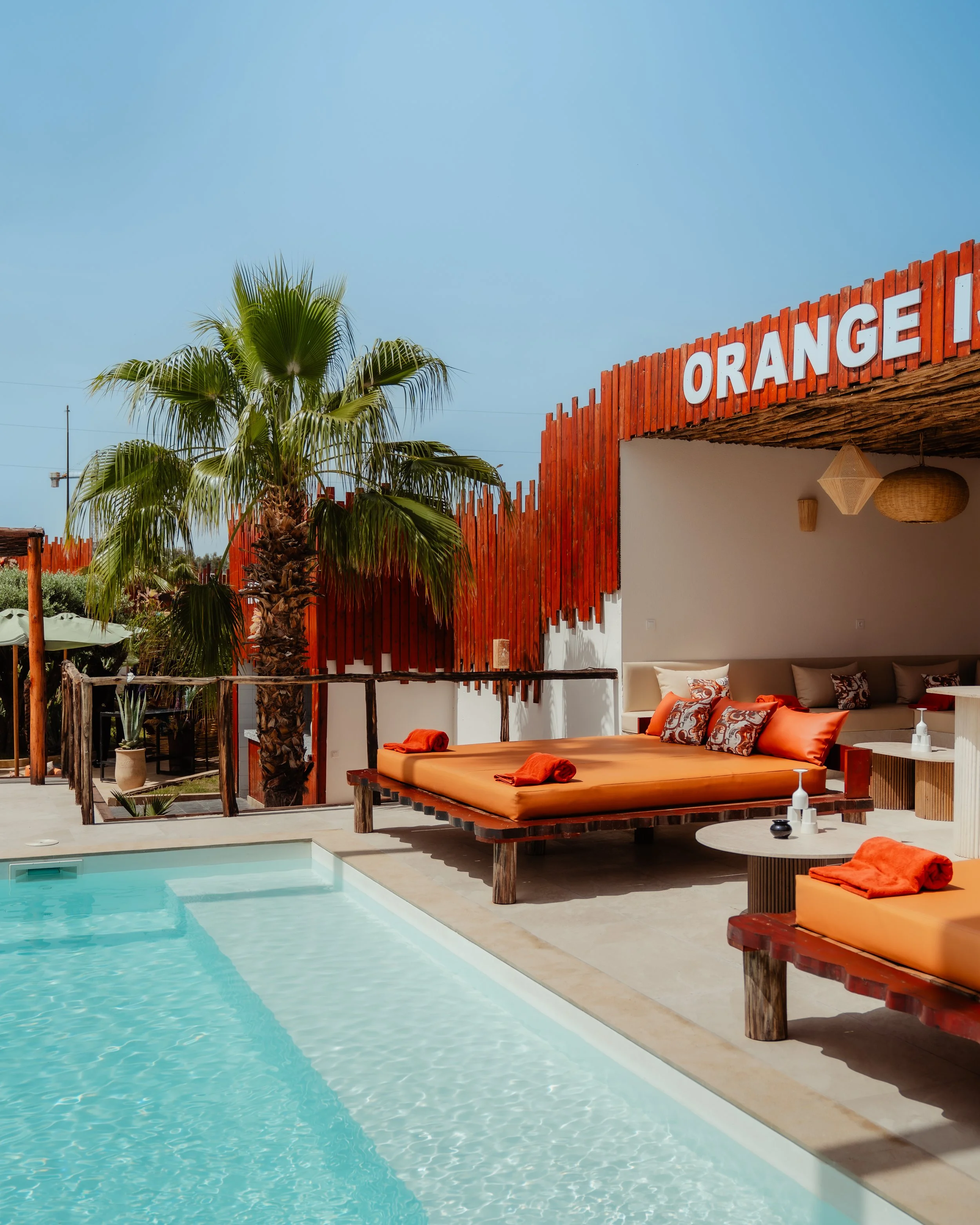 Piscine extérieure avec meubles de détente en bois orange, coussins et serviettes, palmiers en arrière-plan, ciel clair, ambiance chaleureuse et relaxante.