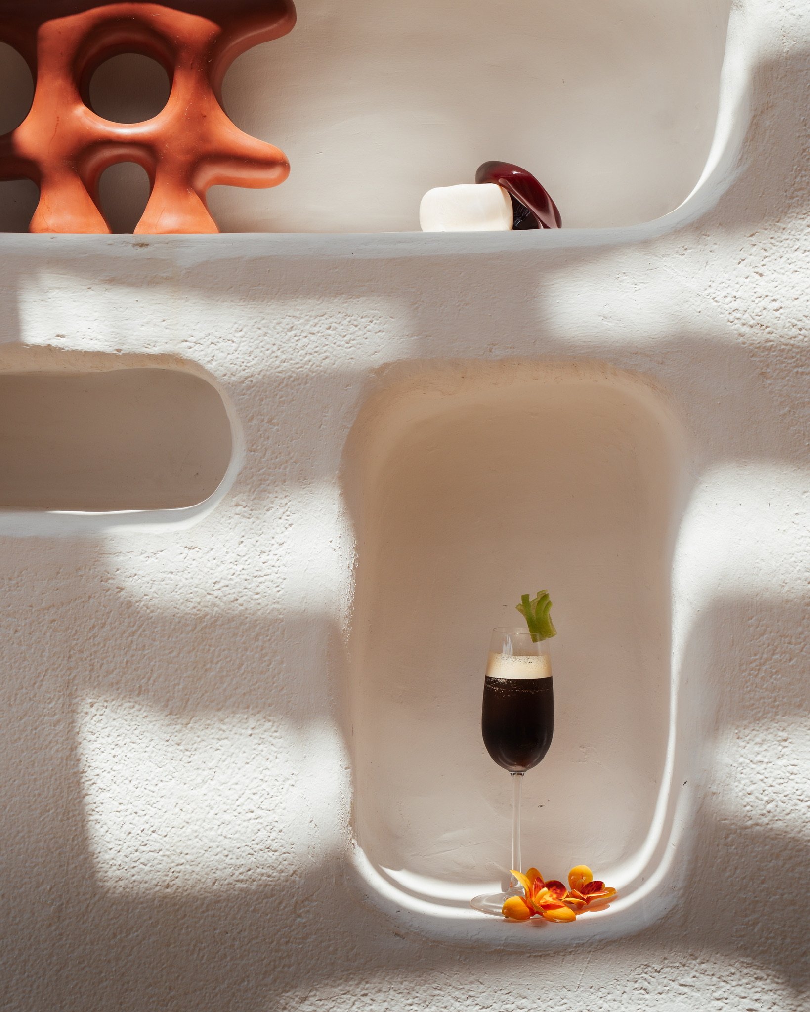 Rangement en forme de niche avec une décoration comprenant un pot en terre cuite, une bouteille de vin avec un verre rempli de vin rouge, une fleur jaune avec des pétales orange, et quelques pétales dispersés.
