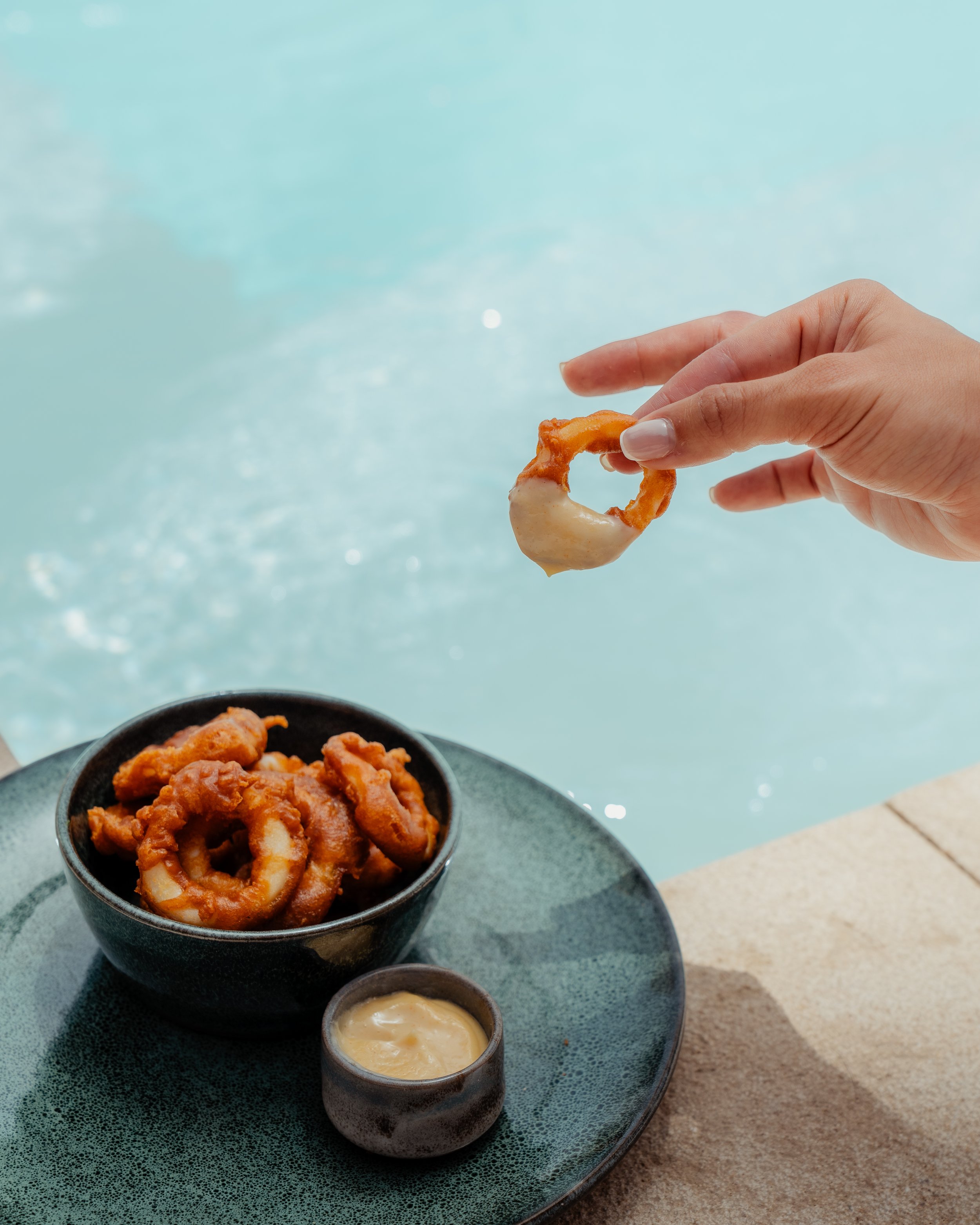 Une main tient une rondelle de calamar frit, avec un bol contenant plus de calamars frits et deux coupes en céramique avec des sauces, le tout sur une assiette en pierre près de la piscine.