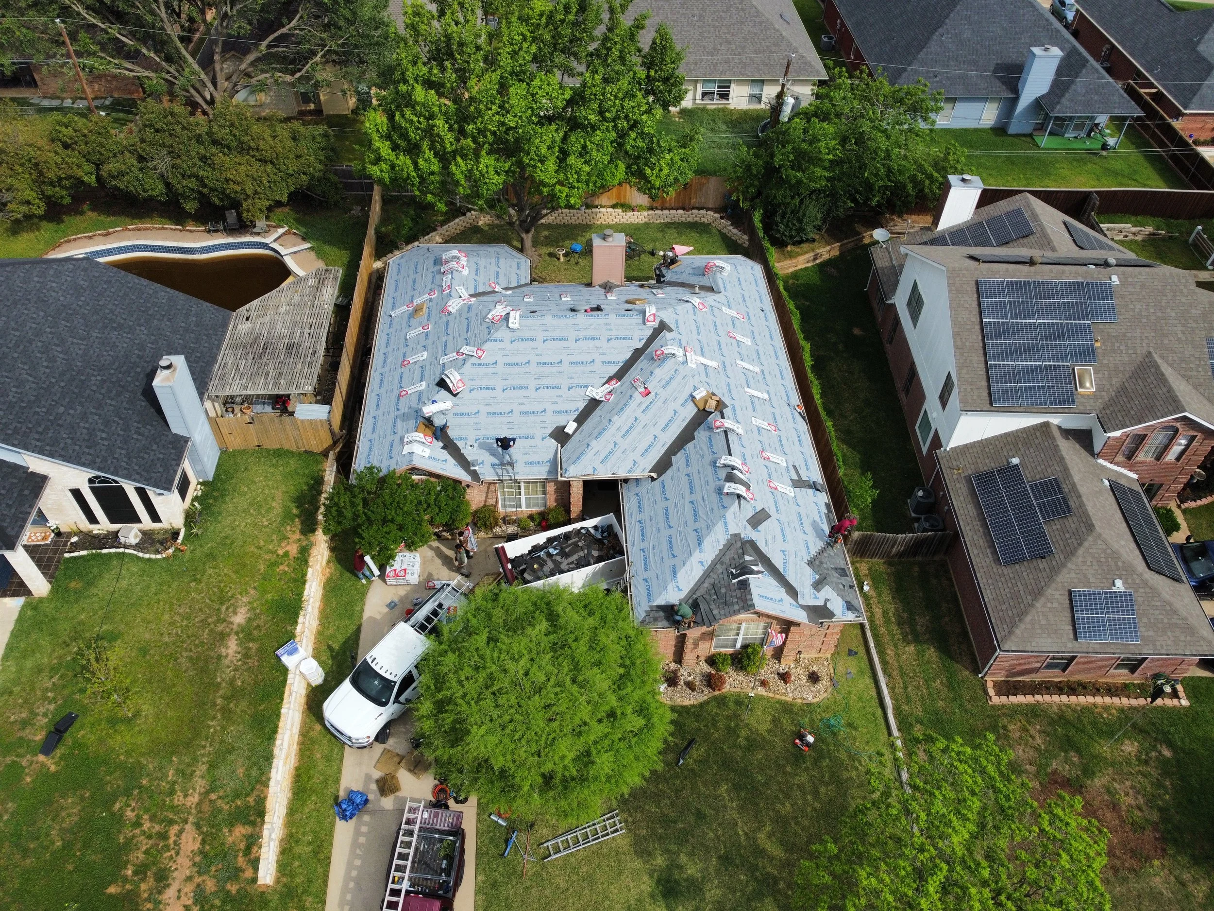 An aerial view of a residential backyard with a house under renovation, showing roofing materials, workers, and tools. Surrounding the house are green trees, neighboring houses, a driveway, and part of a swimming pool.