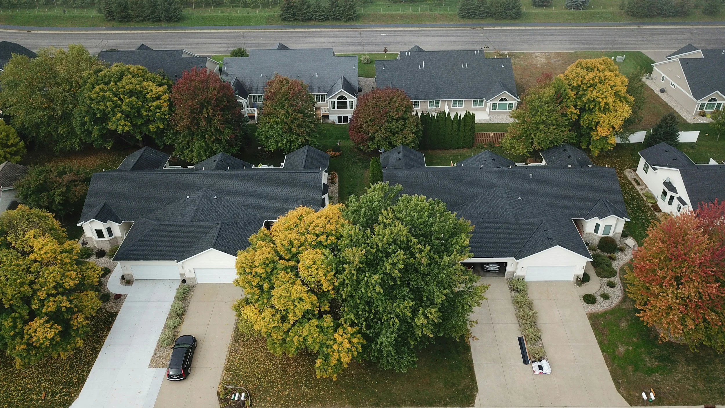 An aerial view of a neighborhood with several houses featuring dark roofs and white or light-colored exteriors. The neighborhood is lined with large, colorful trees with green, yellow, and red leaves, indicating autumn. Streets and driveways are visible, with a couple of parked cars.