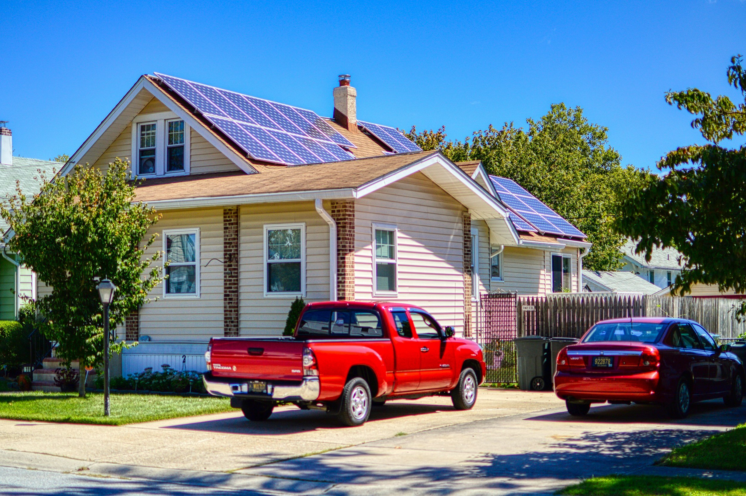 A house with solar panels on the roof, a small front yard with a tree, parked red truck, and other cars in a residential neighborhood under a clear blue sky.
