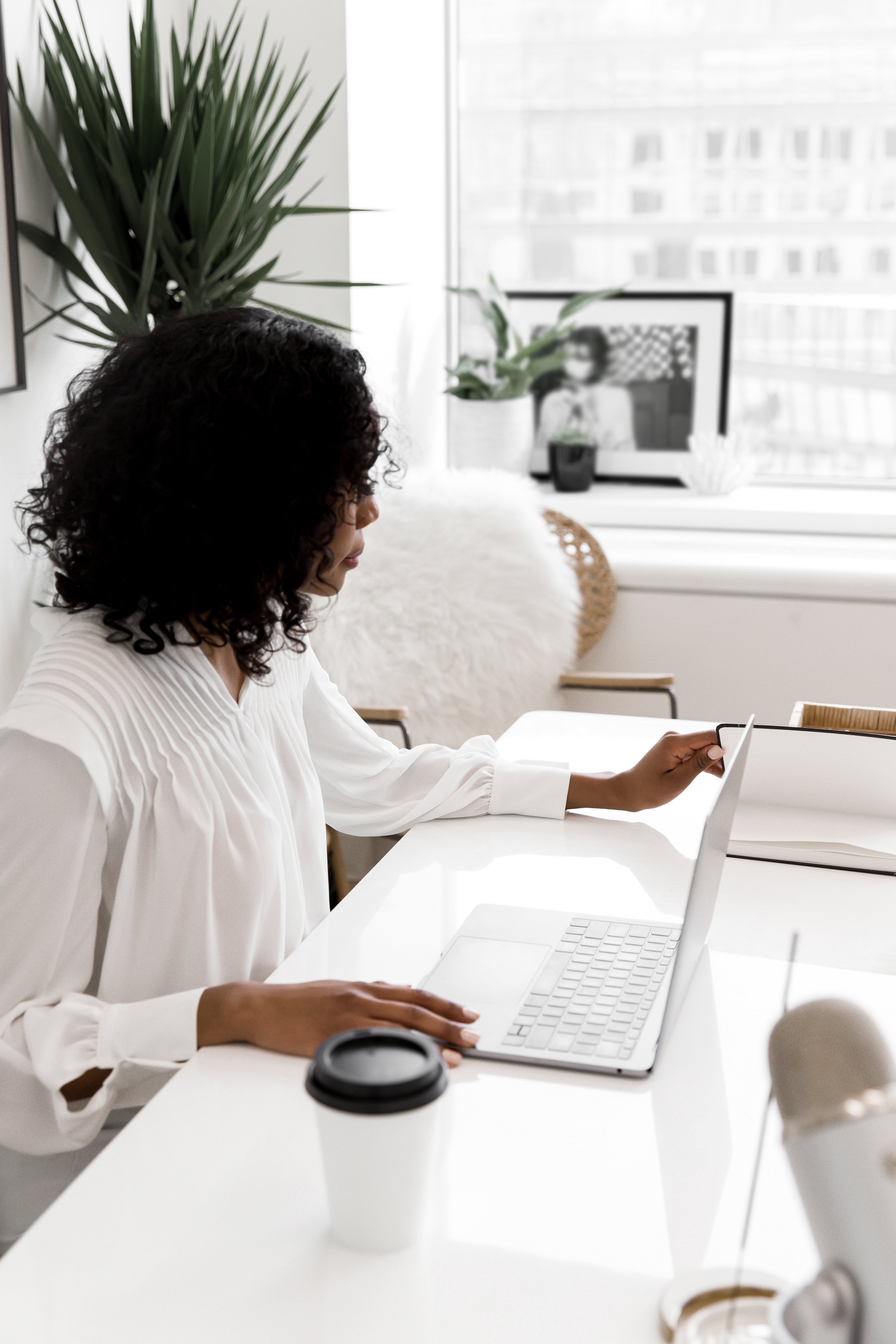 A woman with curly black hair wearing a white blouse sitting at a white desk working on a laptop, with a coffee cup and other items on the desk, in a bright office with large windows, plants, and framed pictures.