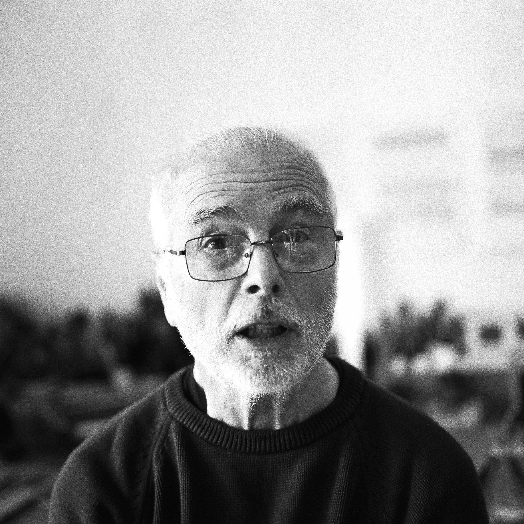 Close-up black and white photo of an older man's face, wearing glasses, with a background blurred out.