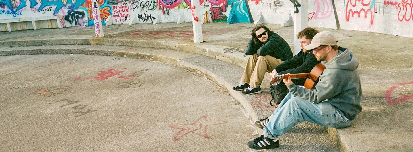 Three young men sitting on a concrete step; one playing guitar, others watching, graffiti-covered wall in background.