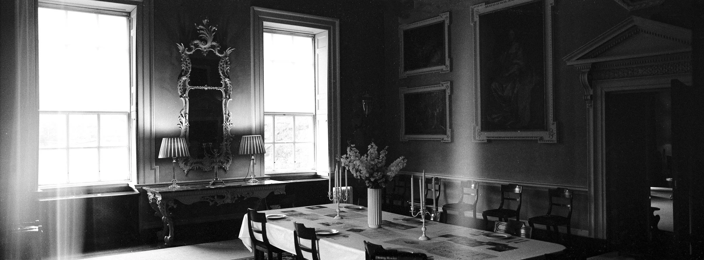 A black and white photo of a vintage dining room with a large table set with a tablecloth and candelabras. There is a large ornate mirror on the wall between two tall windows with light curtains, flanked by two lamps. A vase with flowers is on the ta