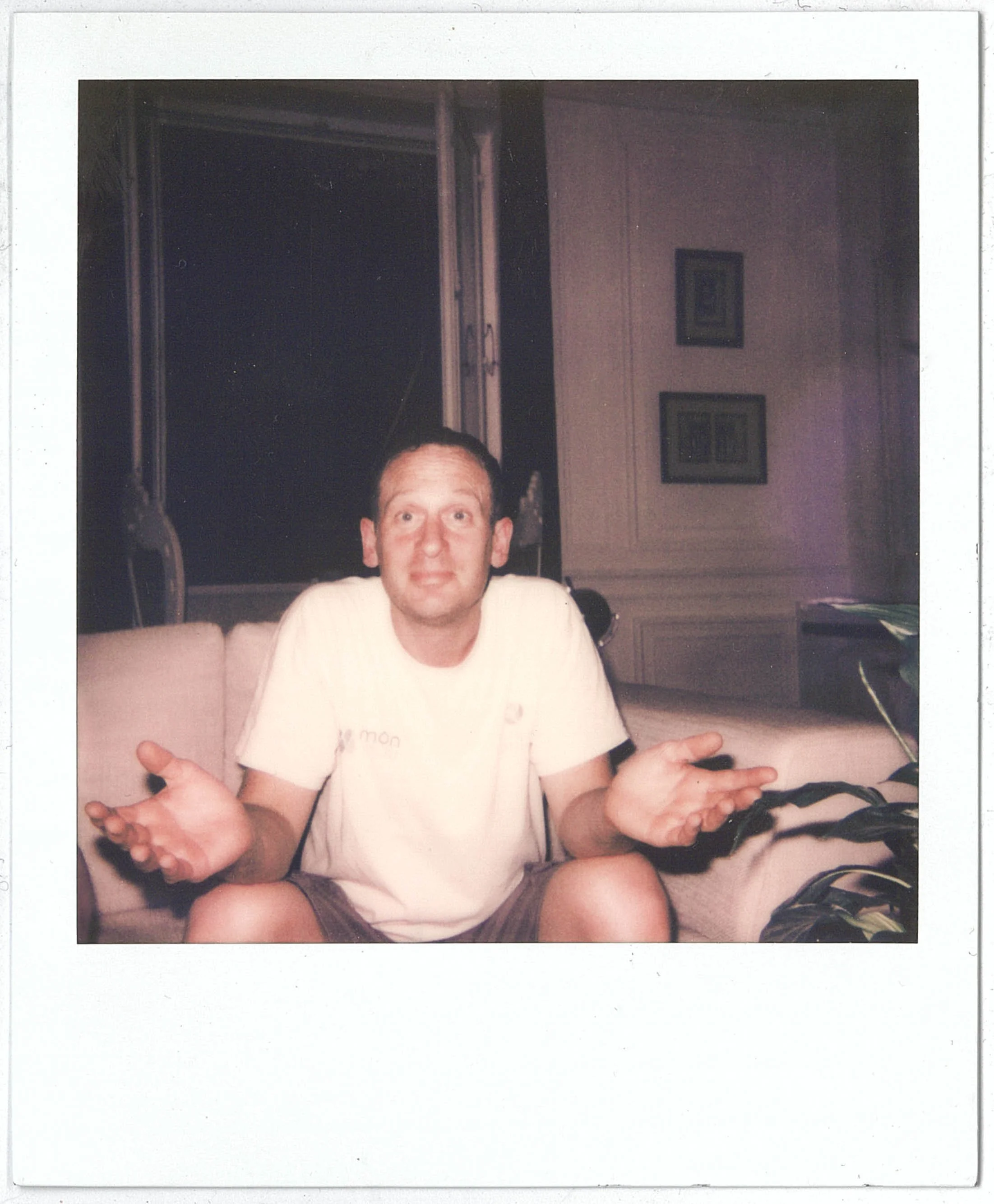 A Polaroid of a man with a puzzled or confused expression, sitting on the floor with arms open and palms facing upward, in a room with wall art and a desk.