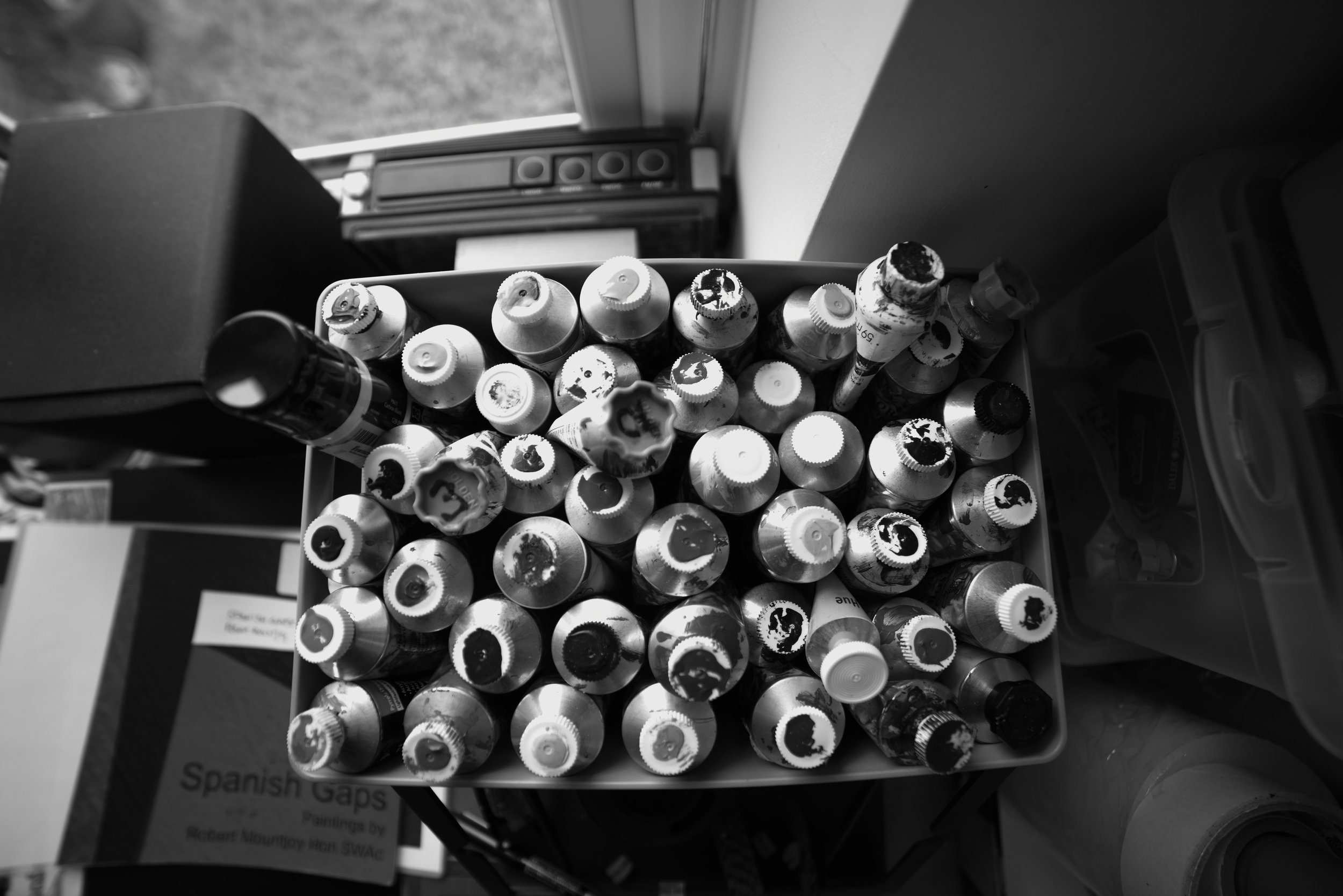Top view of a container filled with used paint bottles with paint remnants on their caps, placed on a table near a window, with a Spanish language book partially visible underneath.
