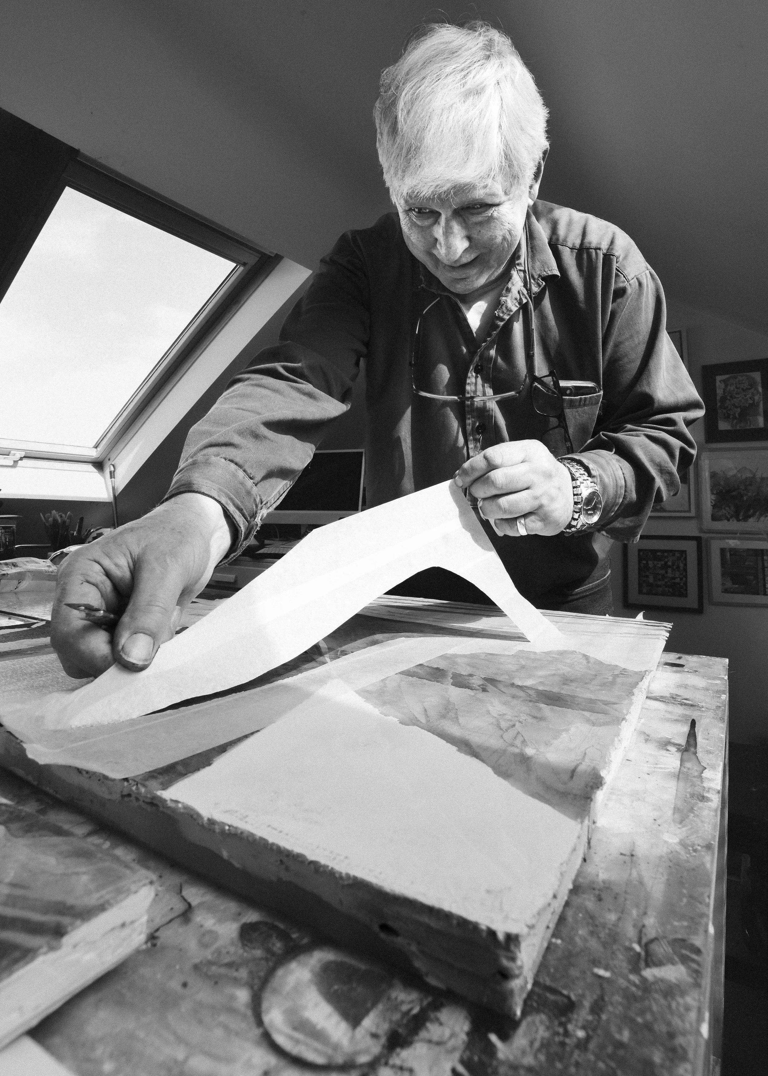 A man with white hair and a ring on his finger is working on a project involving a large piece of wood, possibly carving or shaping it. He is leaning over the work, and the scene appears to be in an artist's or workshop's studio with framed artwork h