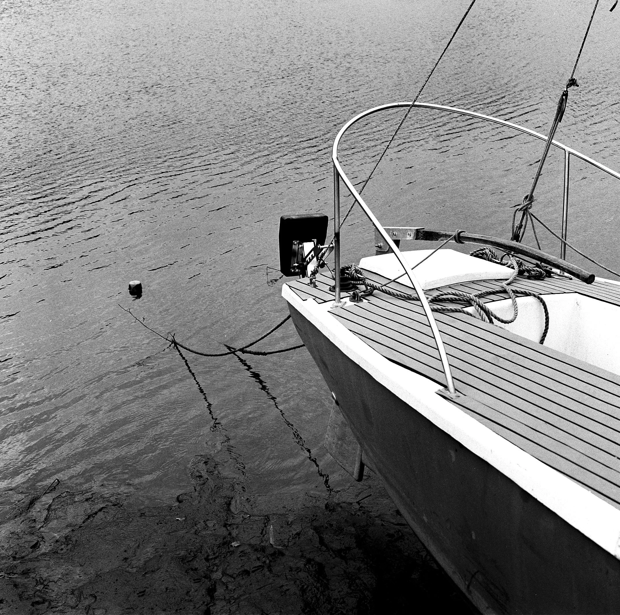 Black and white photo of a boat's bow on a body of water, with visible ropes, railing, and a background of water with gentle ripples.
