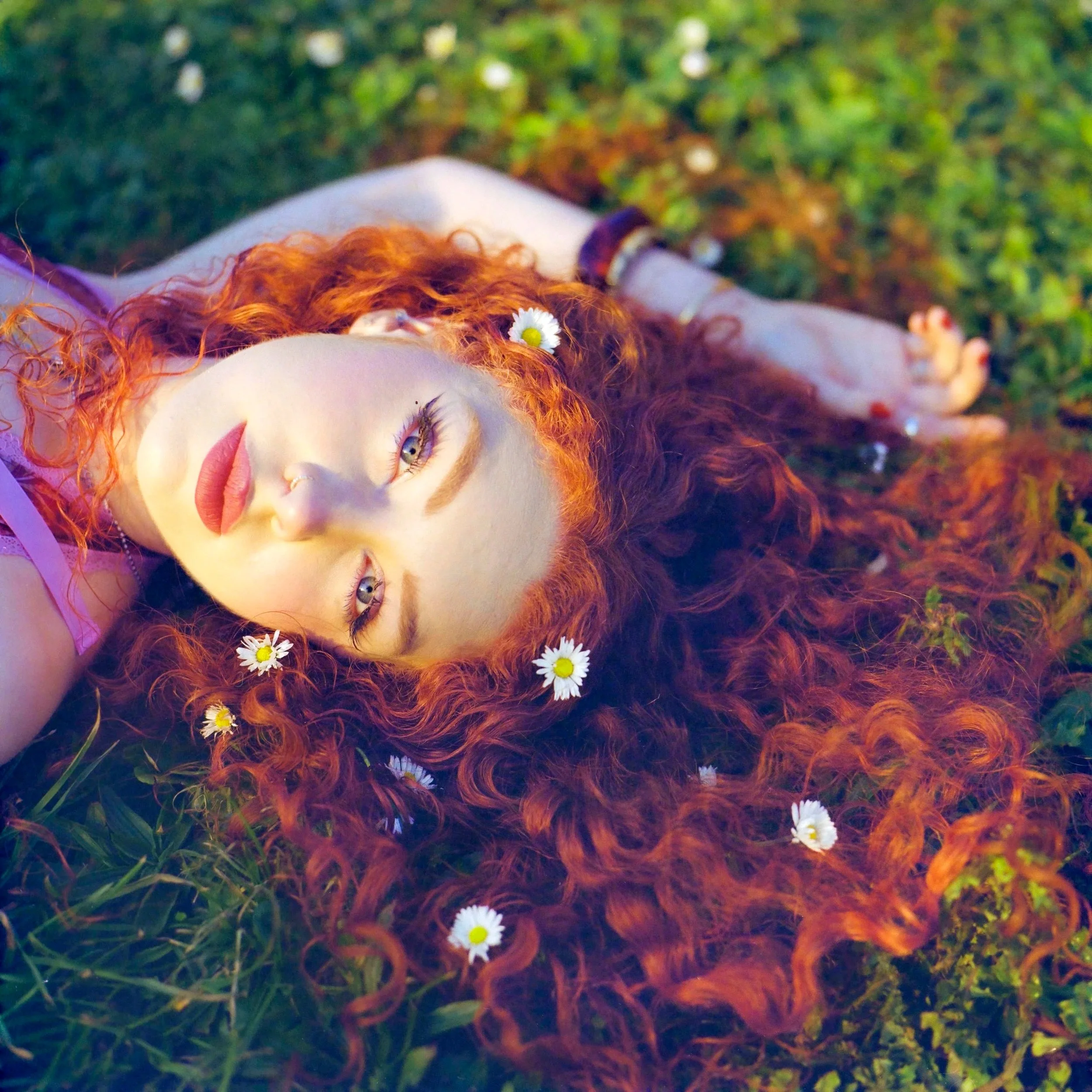 A person with red curly hair lying on the grass, adorned with daisies, with closed eyes and a peaceful expression.