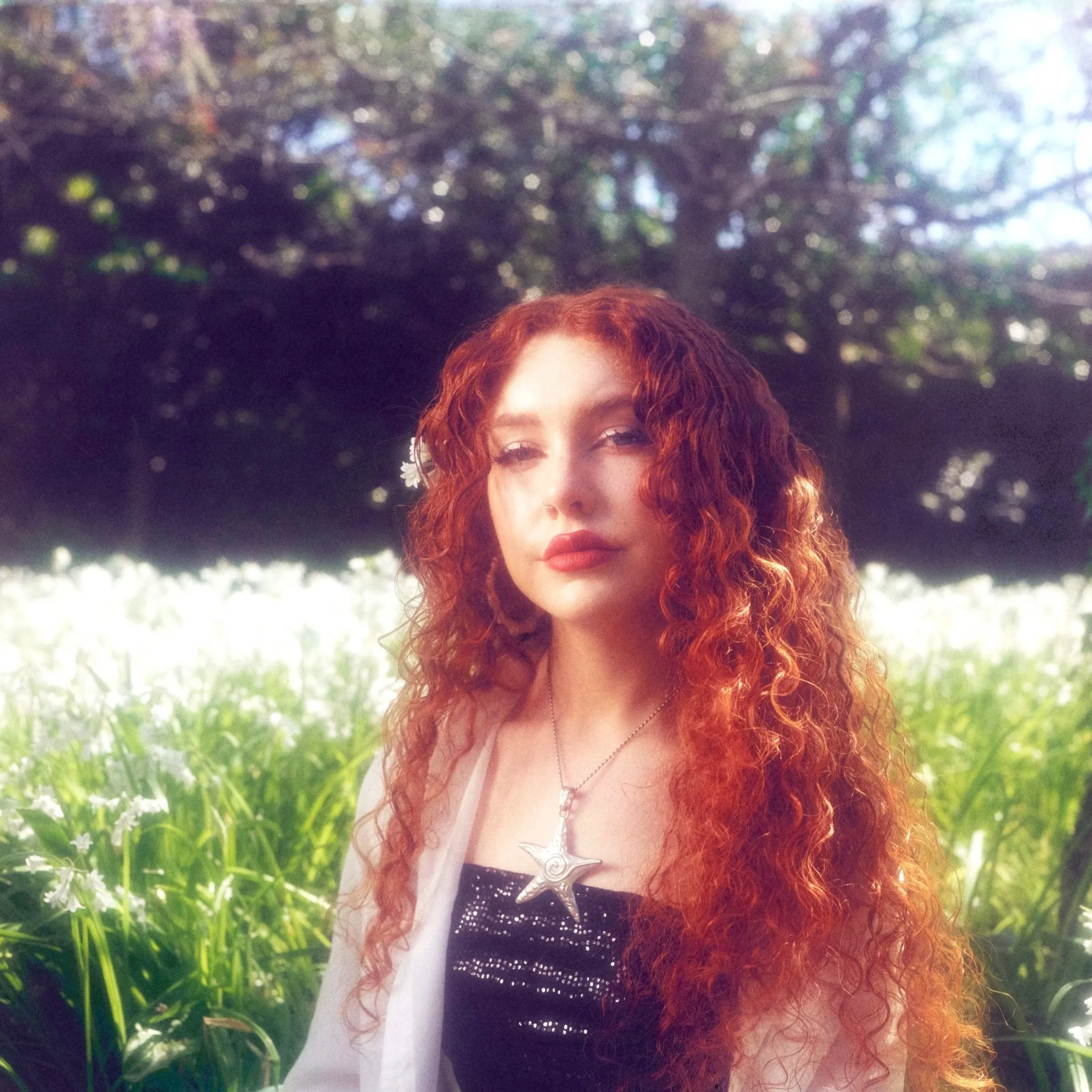 Part of a woman's face with red curly hair, wearing a silver star necklace, in front of a background of greenery and blurred trees.