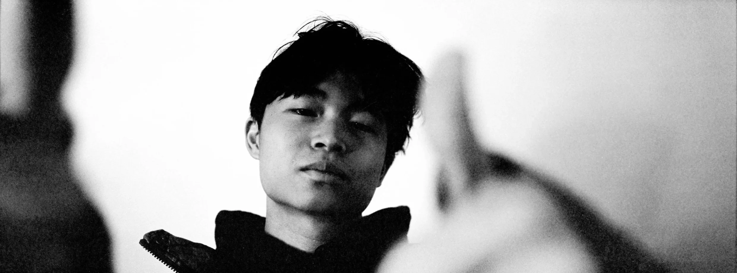 Black and white portrait of a young man with dark hair, slightly tilted head, wearing a dark jacket with a textured collar, looking at the camera with a subtle expression.