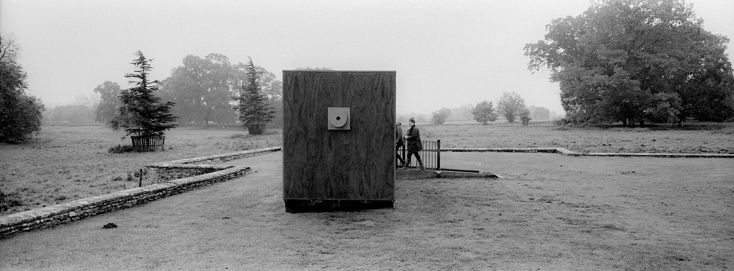 A black and white photo of a park with a large rectangular wooden structure with a small square attached to it, and two people walking behind it near a gate. There are trees and shrubs in the background.
