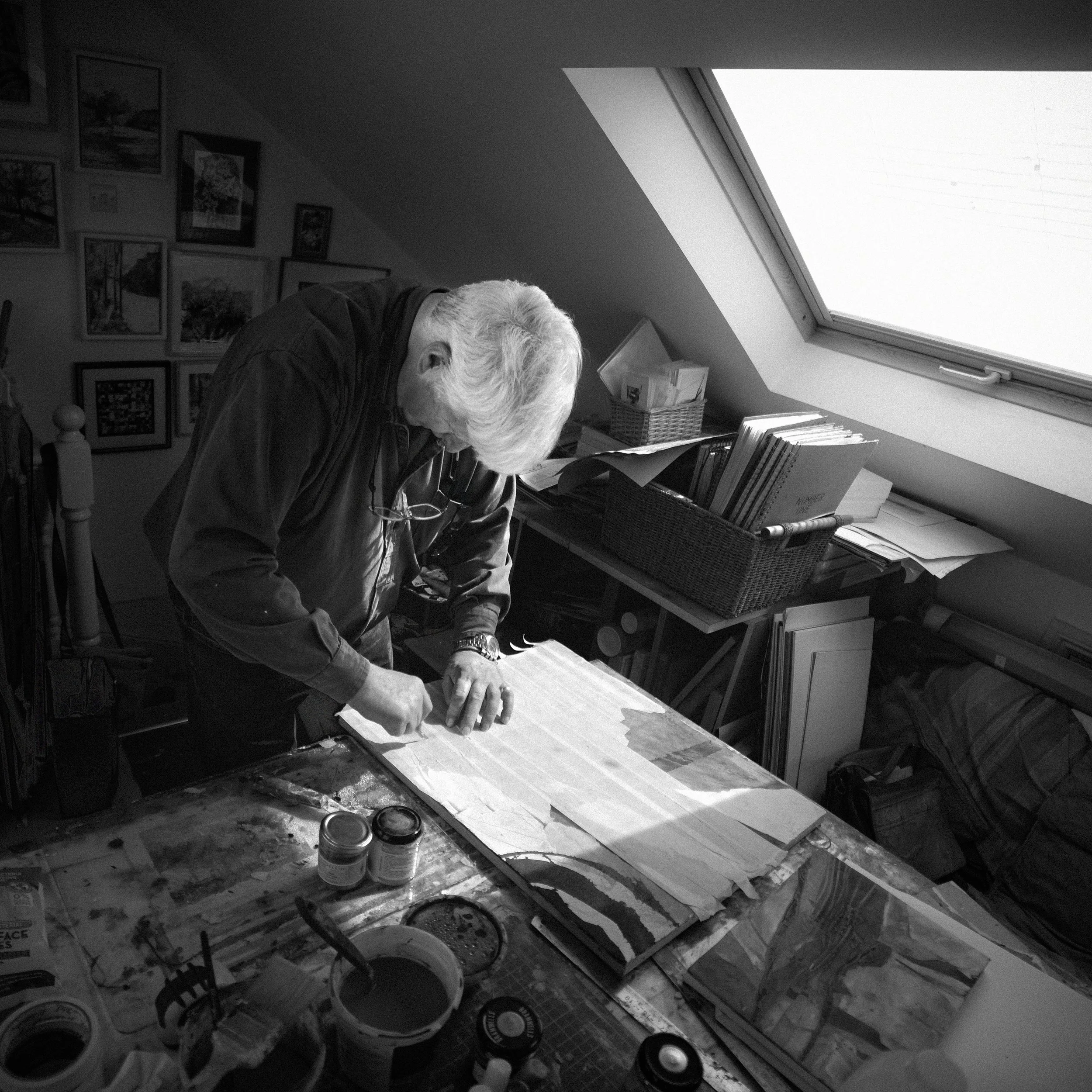 An elderly person with white hair working on a craft project in a cozy room with art on the walls and various supplies on a table.