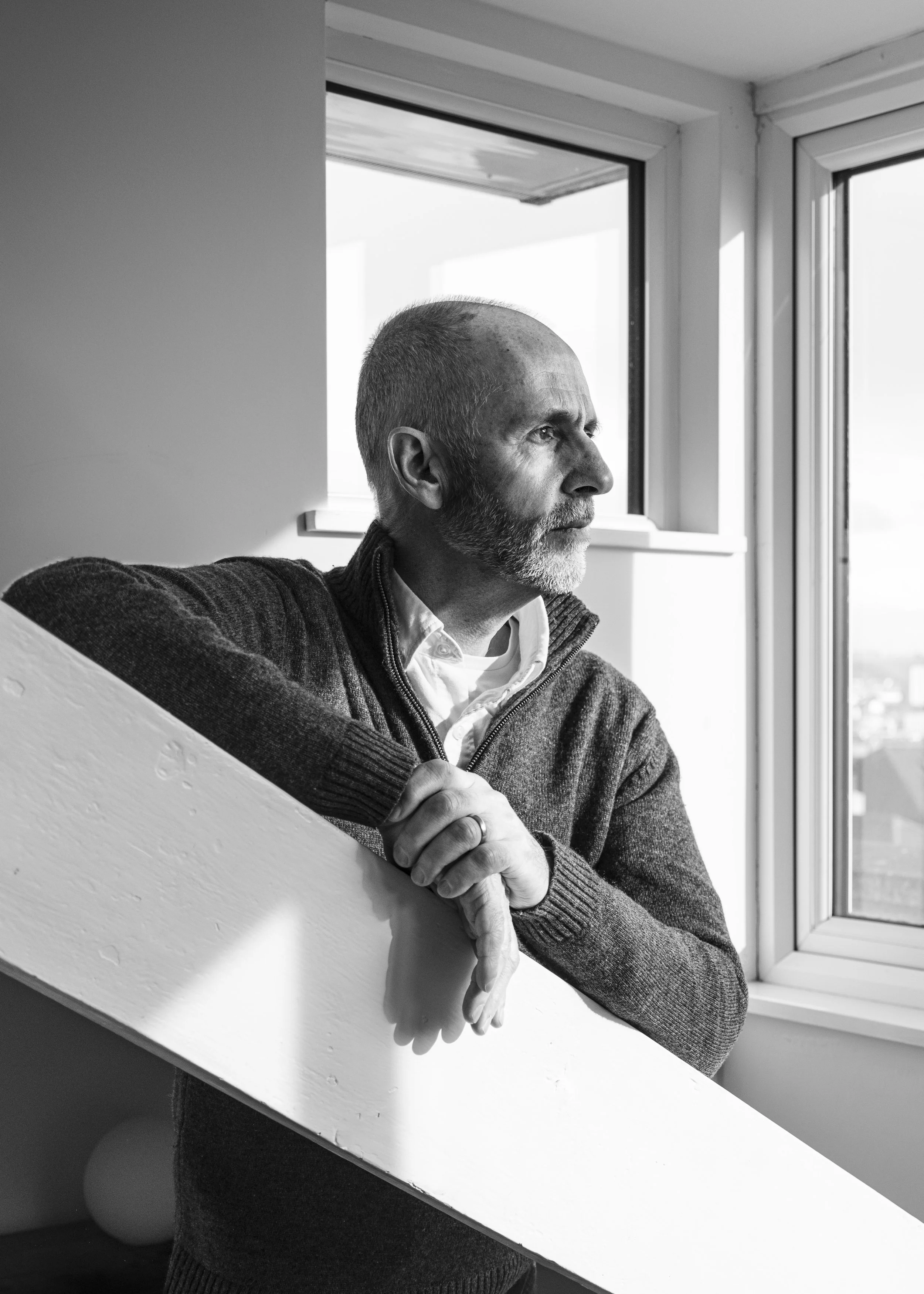 Black and white photo of a middle-aged man with short hair and beard, sitting near a window, looking thoughtfully outside, wearing a sweater and shirt.