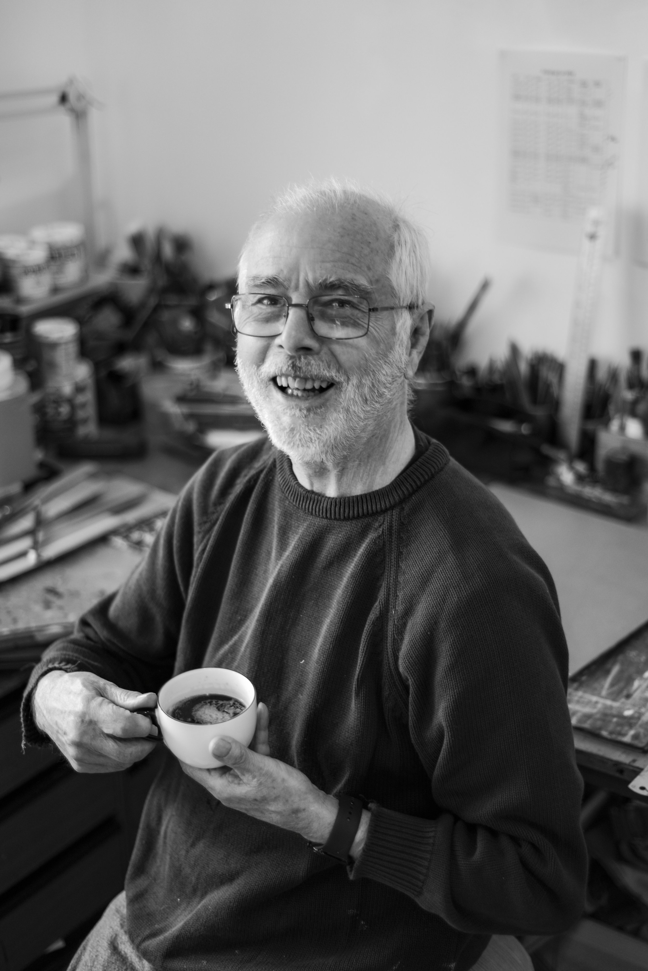 An elderly man with glasses and a beard smiling, holding a cup of coffee in a messy room with shelves of cans and tools.
