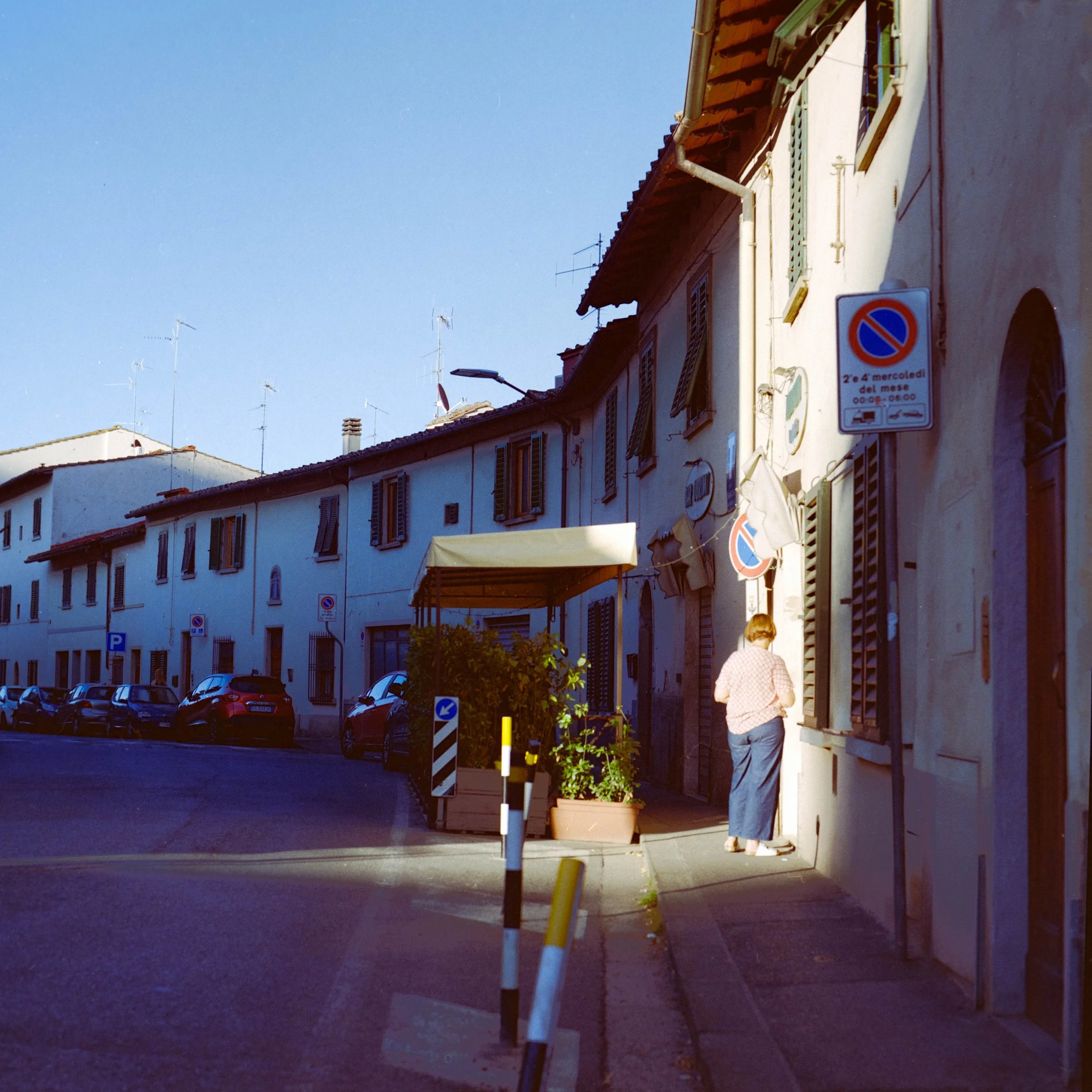 A street scene with parked cars, residential buildings with closed shutters, and a person walking on the sidewalk under a clear sky.