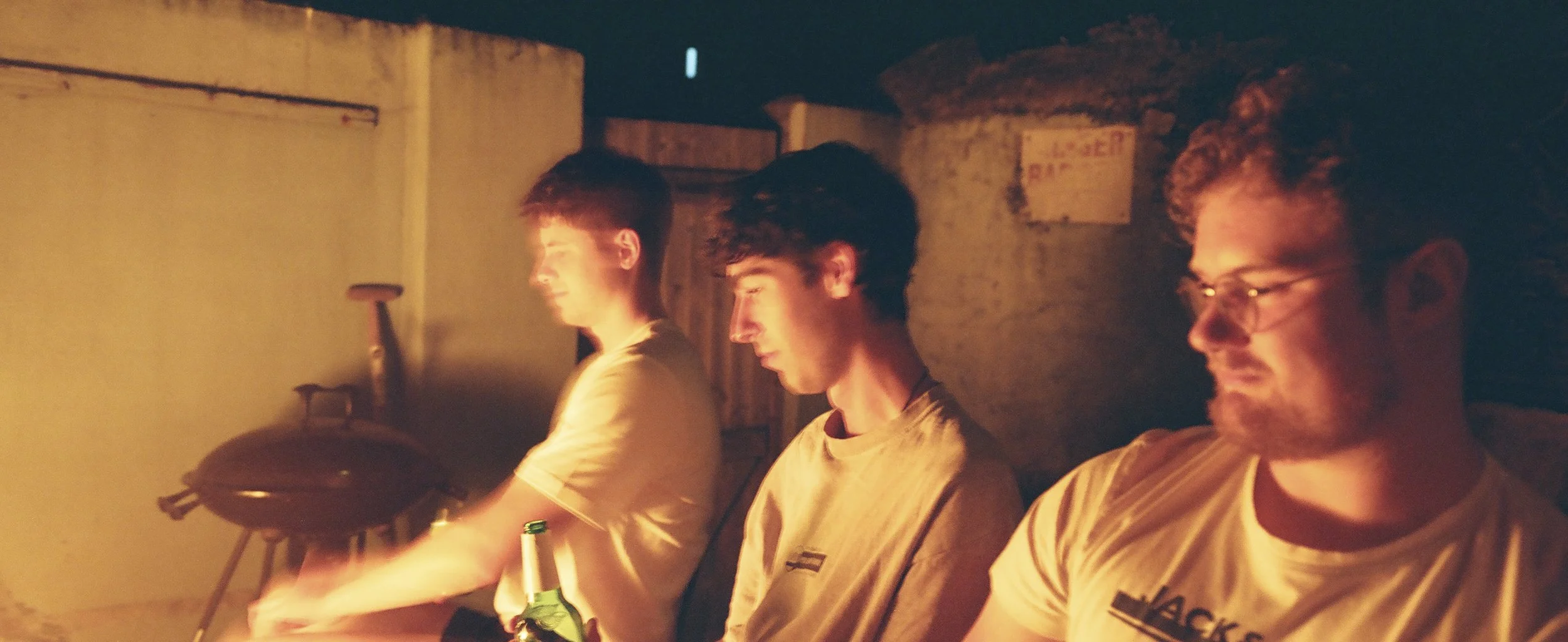 Three young men sitting in a dimly lit room, possibly around a campfire or in a cozy setting, with relaxed and happy expressions.