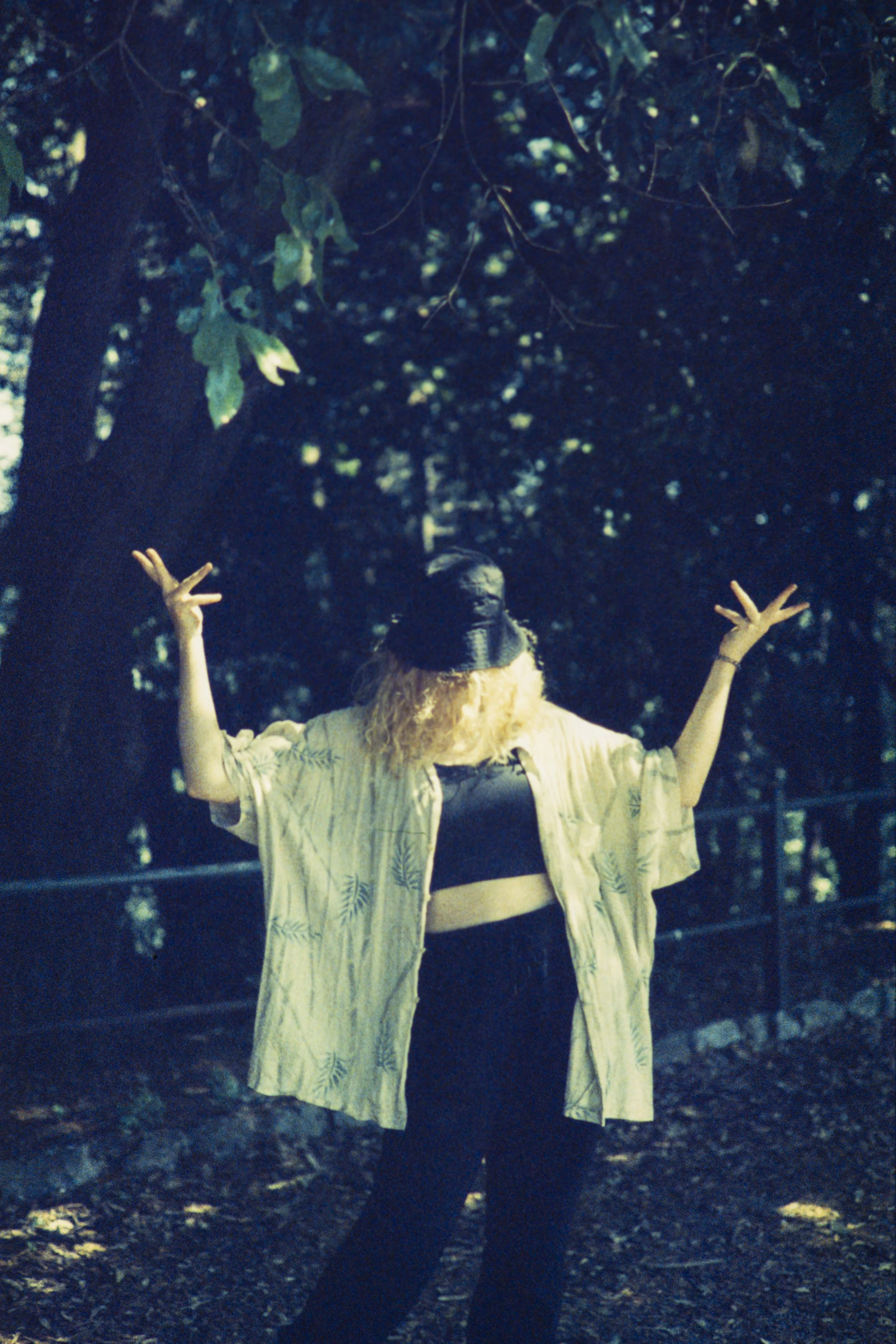 A person with long curly hair wearing a yellow shirt with leaf print, black pants, and a black top underneath, standing outdoors in a wooded area with their arms raised and fingers spread, partially hiding their face.