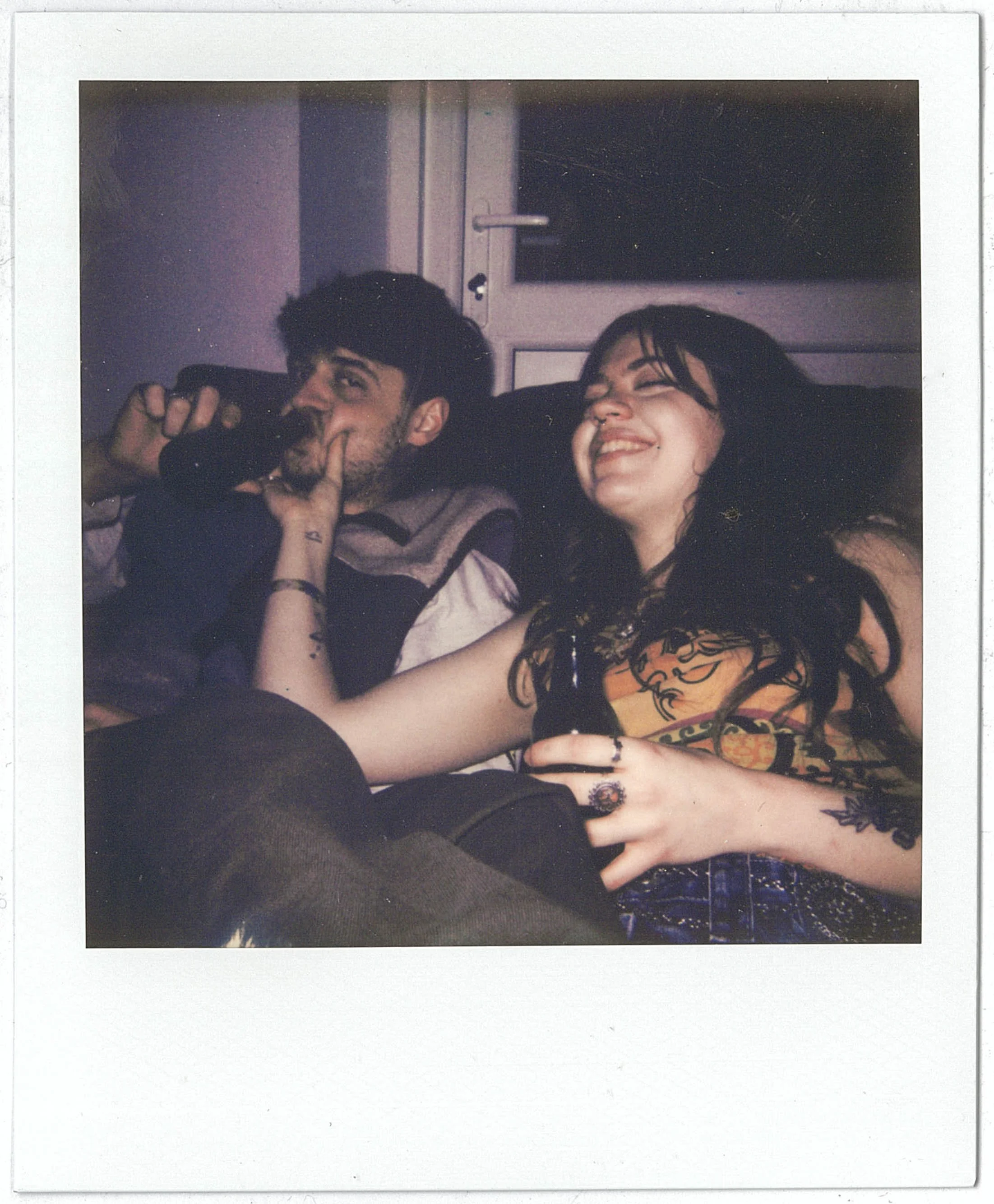 A Polaroid photo of a young man and woman sitting together, smiling and laughing. The man is holding a camera, and the woman has dark hair with bangs and tattoos visible on her arm.