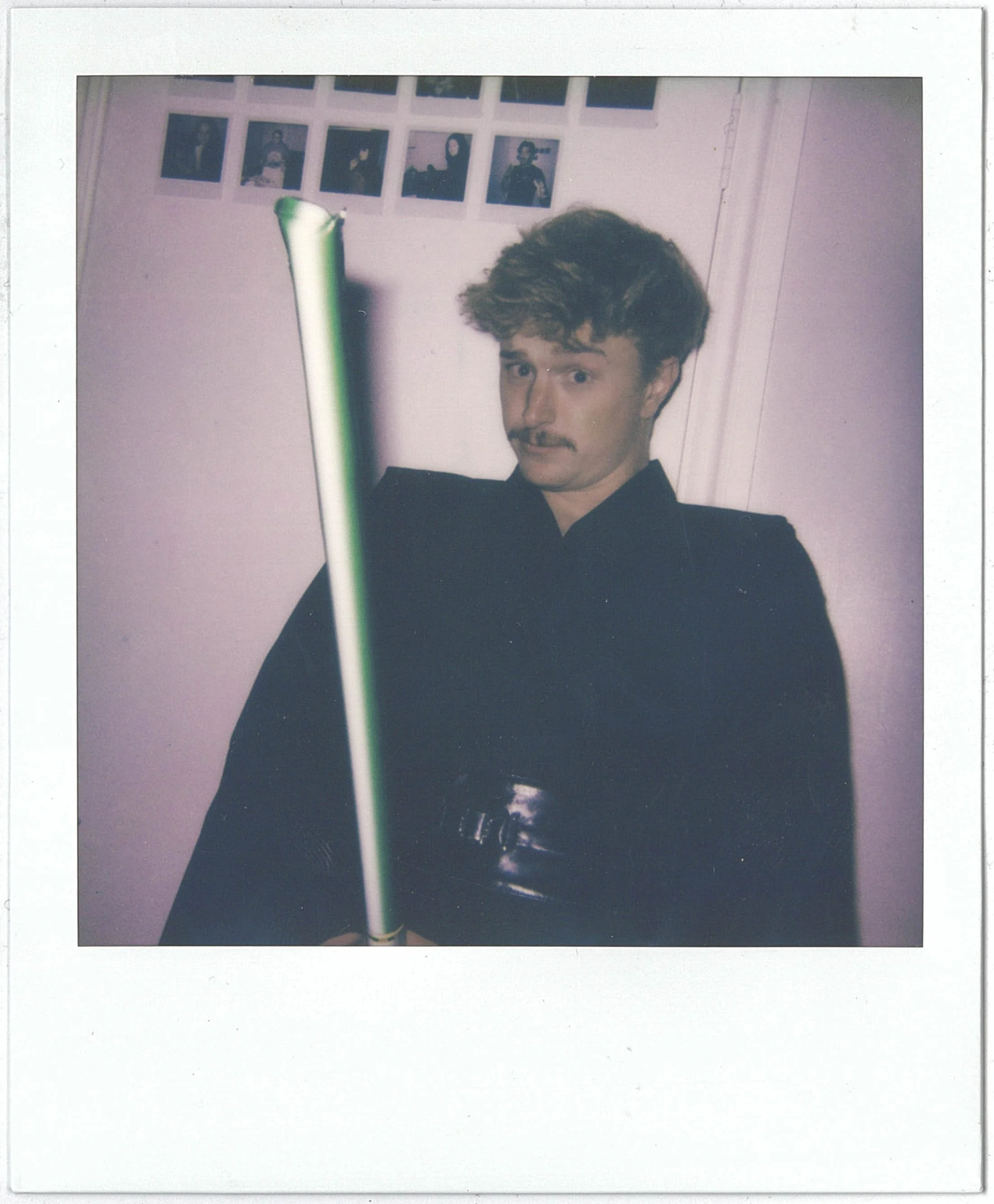 A Polaroid of a young man with curly hair and a moustache, taking a Polaroid photo of himself, holding it up in front of a pink wall with framed photographs.