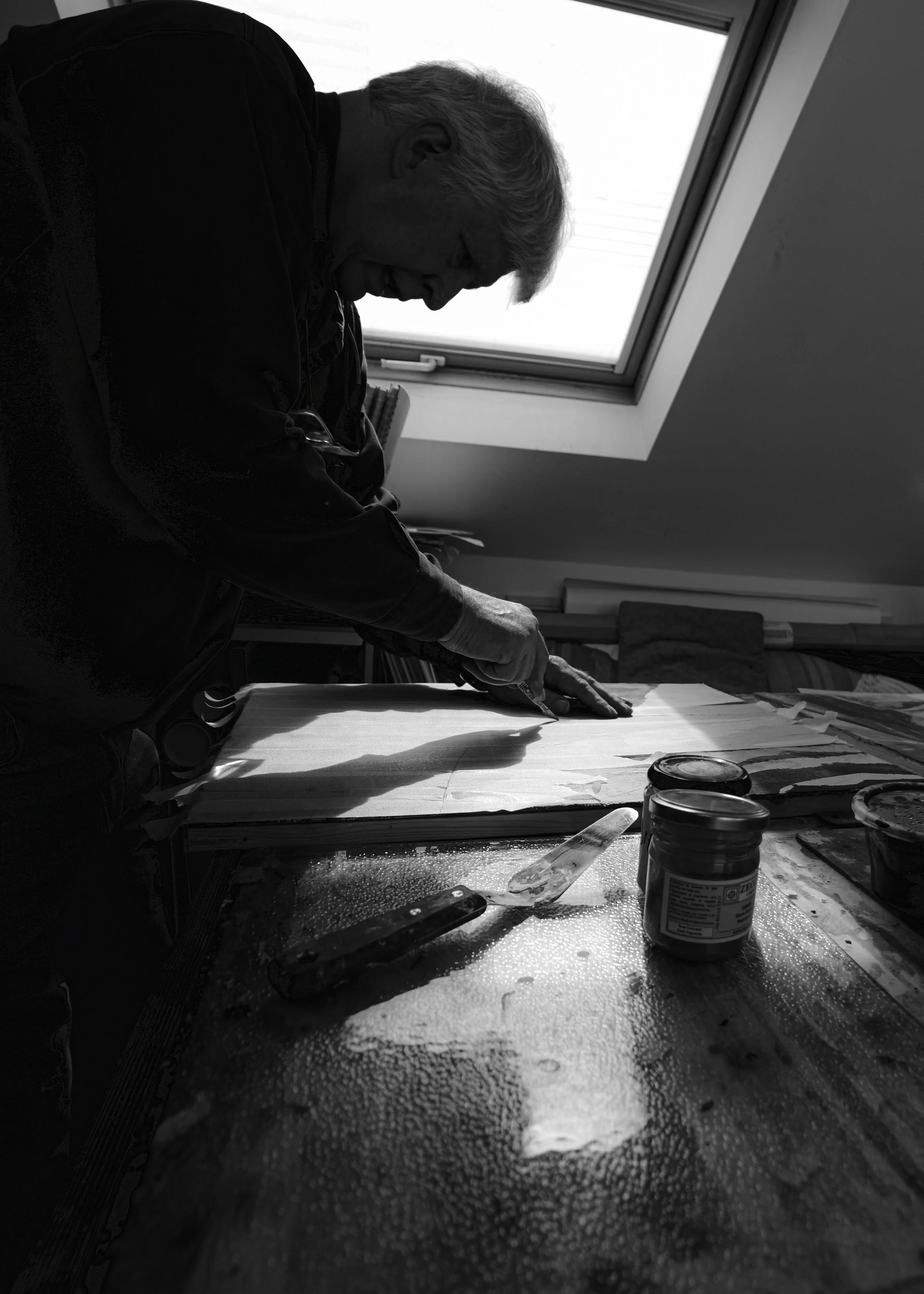 A black-and-white photo of an elderly man with gray hair working at a table near a window. He appears to be focused on a project, possibly painting or drawing, with art supplies and jars on the table.