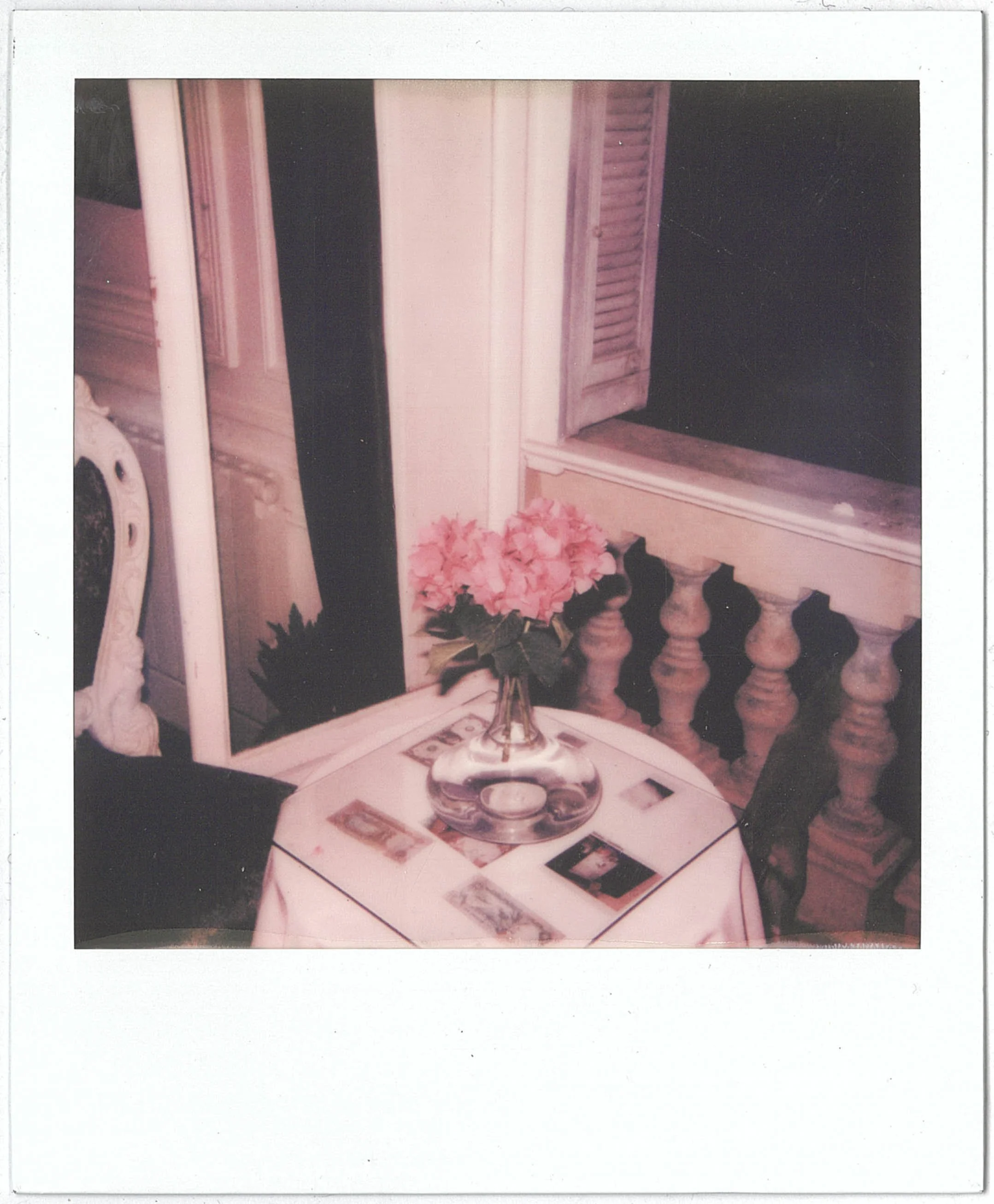 A Polaroid of a small table with a pink cloth, a bouquet of pink flowers in a vase, a few framed photographs, and a decorative urn, positioned against a white balustrade with turned balusters.