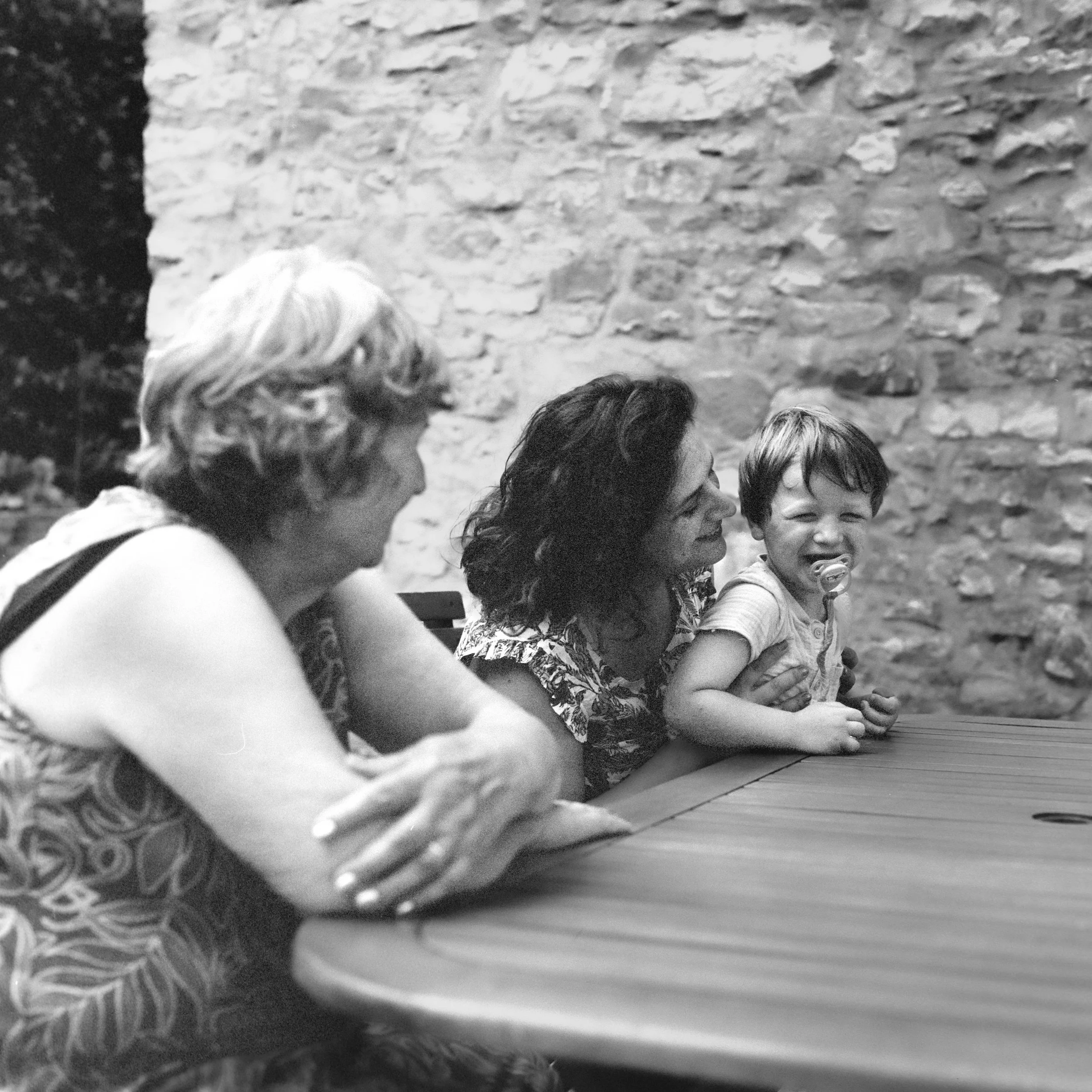 A black and white photograph showing the heads of several children sitting at a table, with a stone wall in the background. The children have various hair styles, and some are smiling.