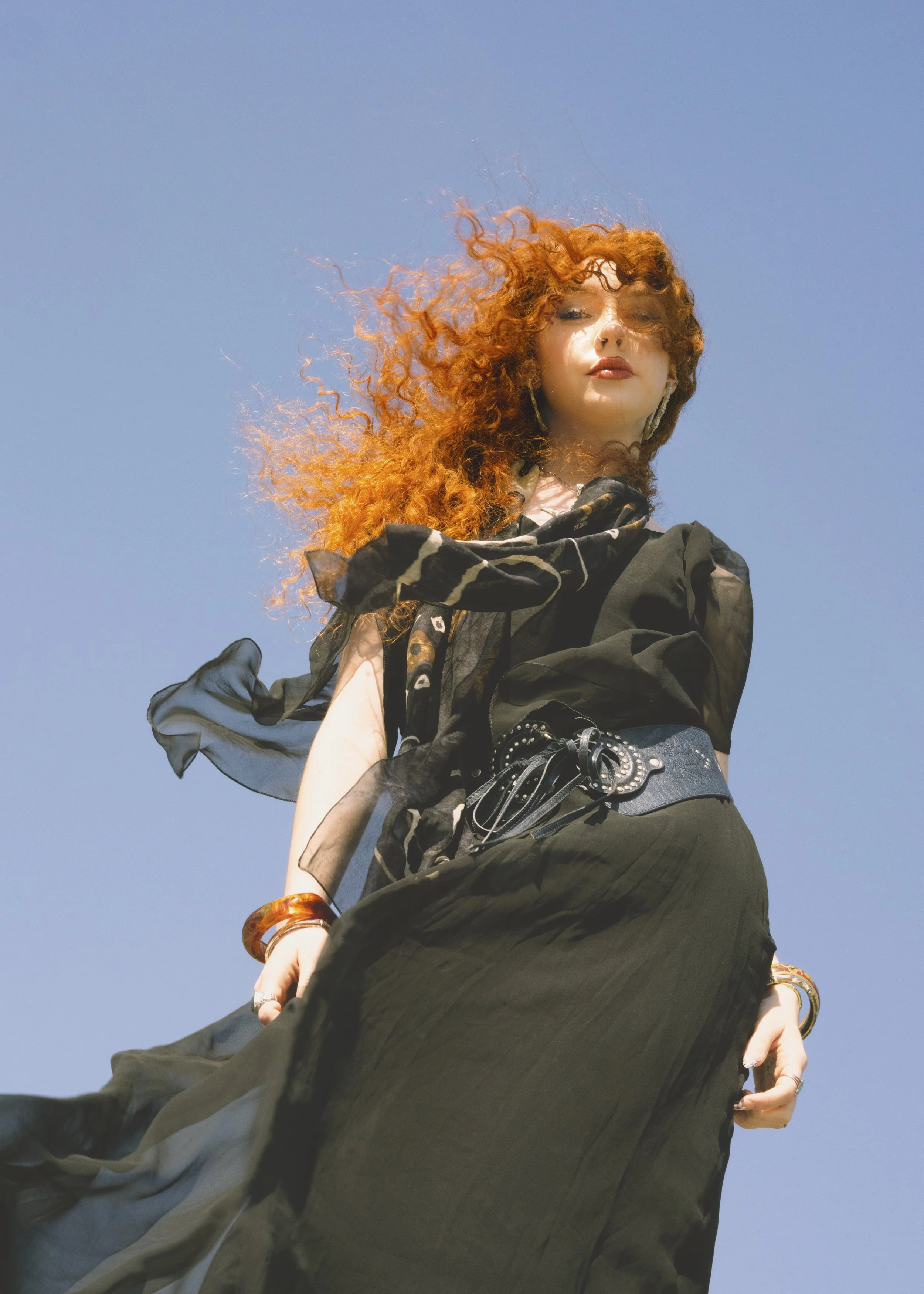 Low-angle view of a woman with curly red hair, wearing a black dress, accessories, and black gloves, standing outdoors against a clear blue sky.