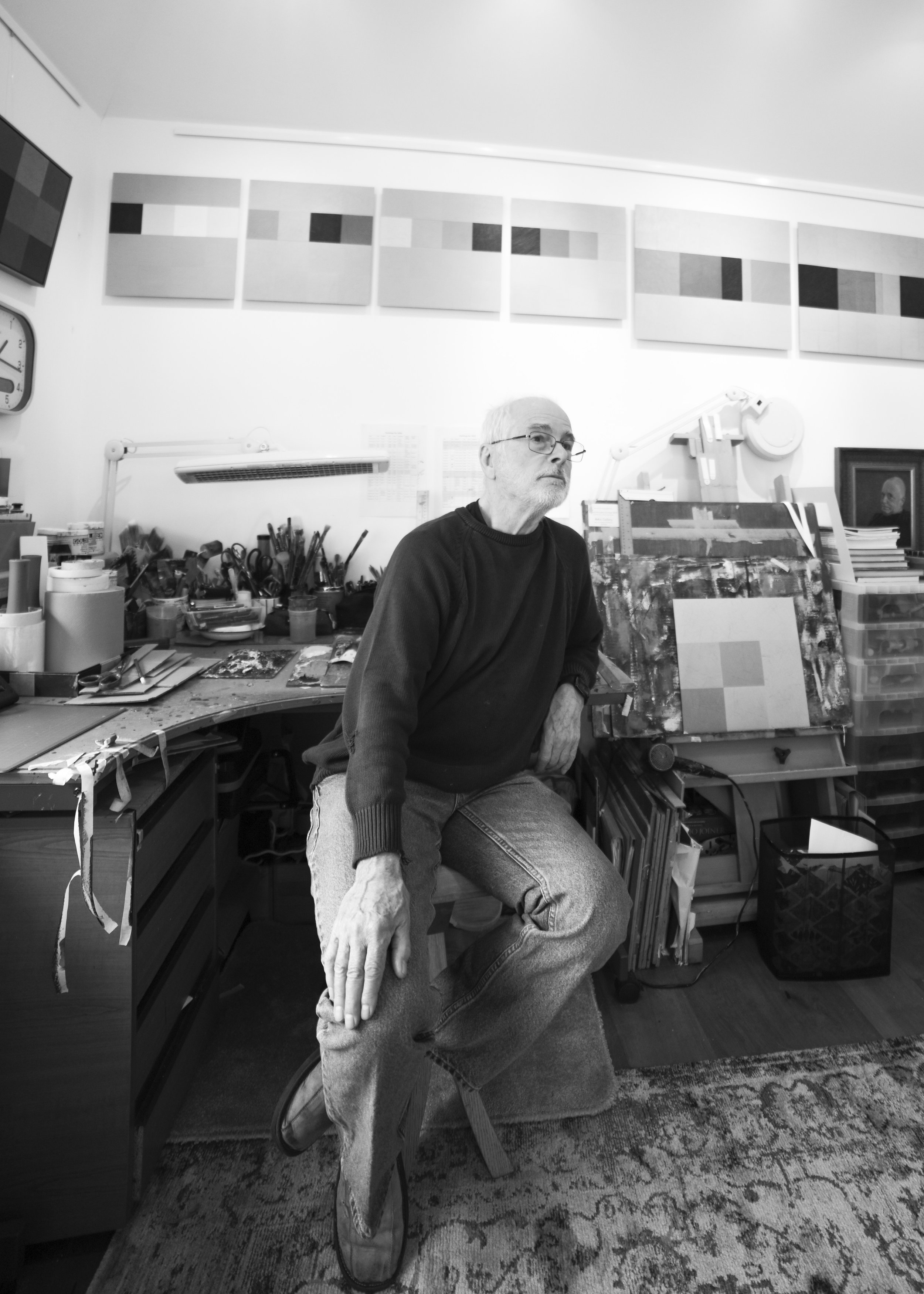 An elderly man with glasses sitting in an art studio surrounded by artwork, art supplies, and framed pictures, with a serious expression.