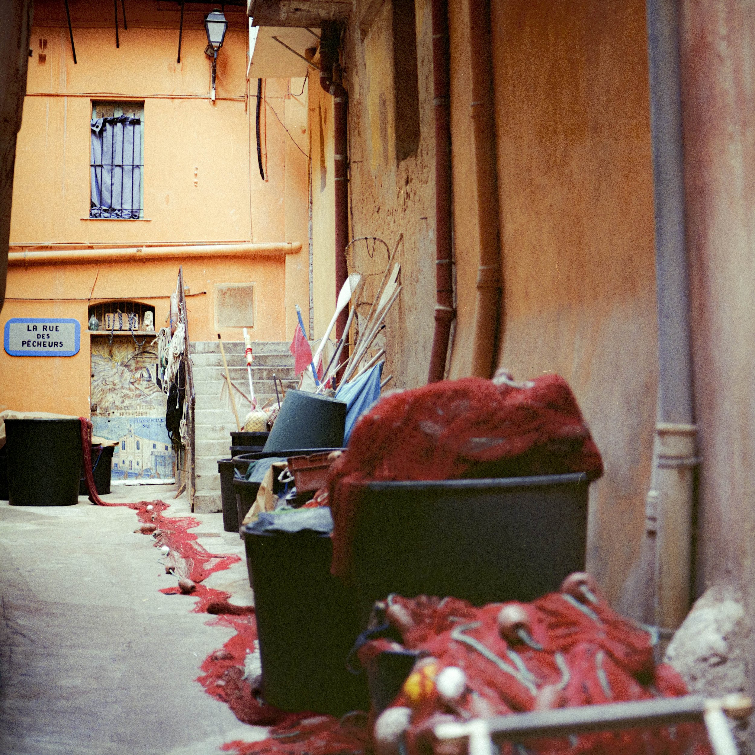 Close-up of a red shoe with a patterned design on the sole, placed in front of a row of black flower pots and a colonial style building with peach-colored walls, windows with metal grilles, and a street sign reading 'LA RUE DES PECHERS'.