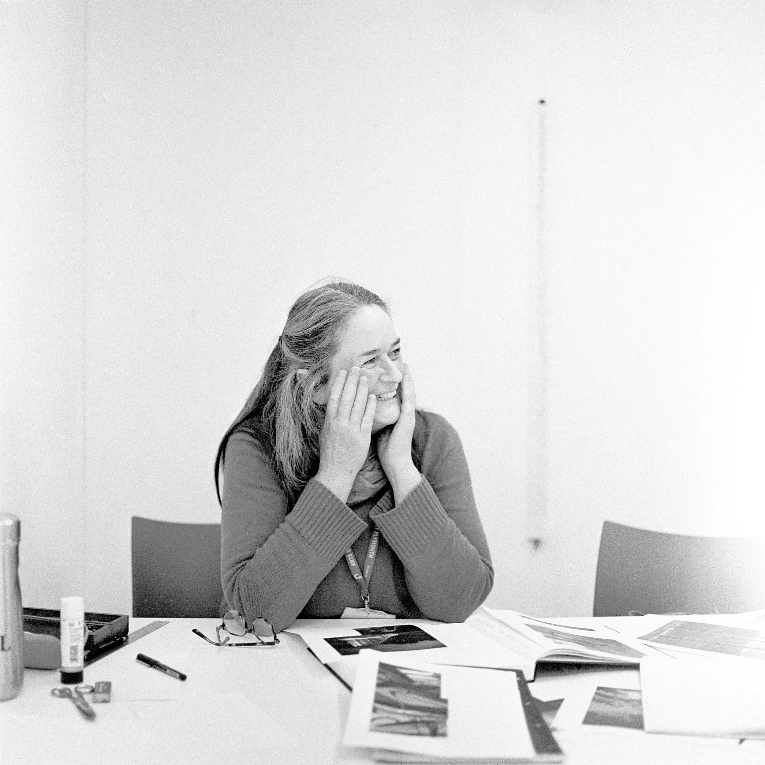 A woman sitting at a desk with her hand on her face, surrounded by papers, photos, and office supplies in a classroom or office setting.