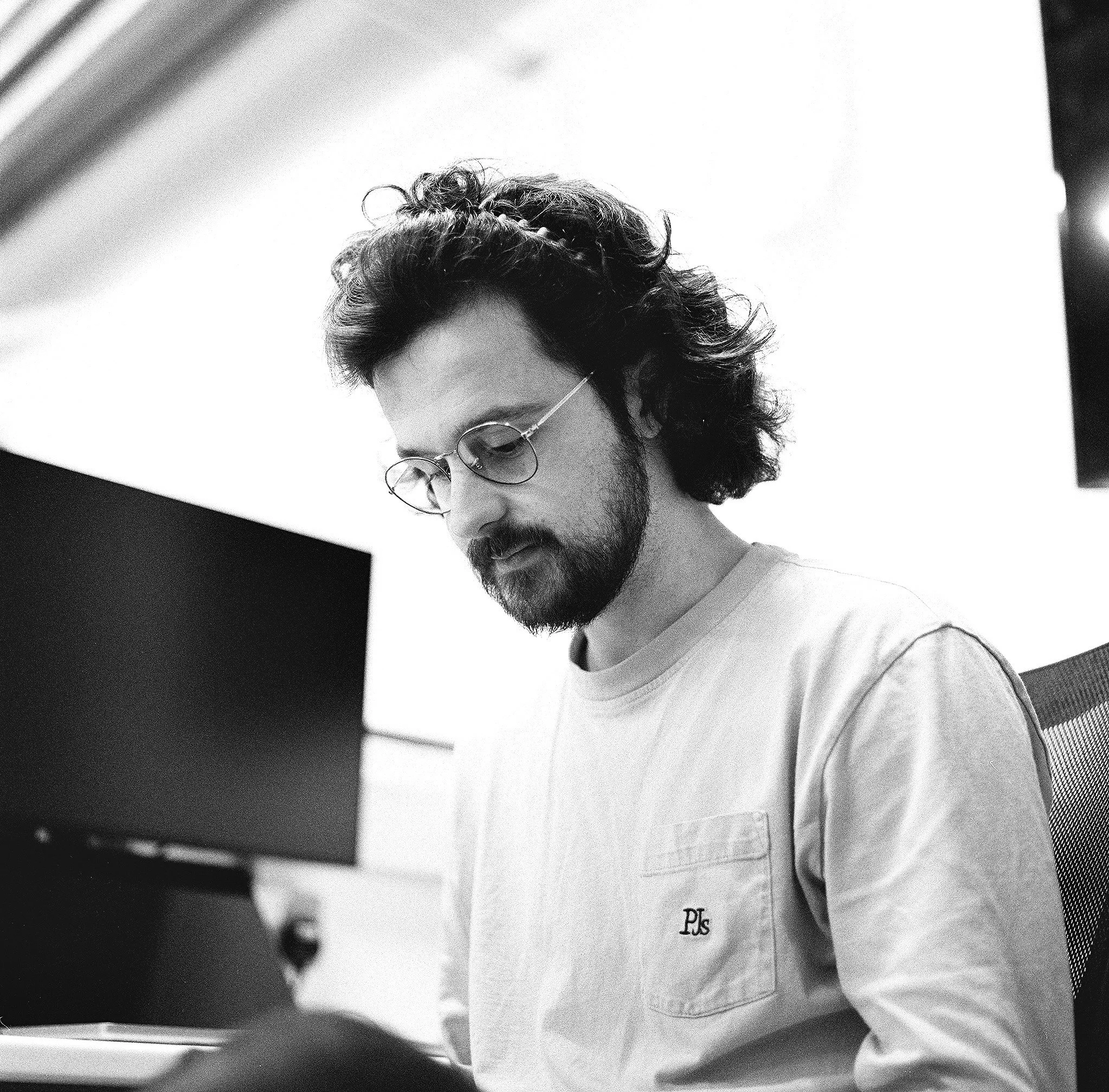 Black and white photo of a man with curly hair and glasses, wearing a white shirt with embroidered initials 'D T' and 'P S' on the pocket, indoors with a spiral staircase in the background.
